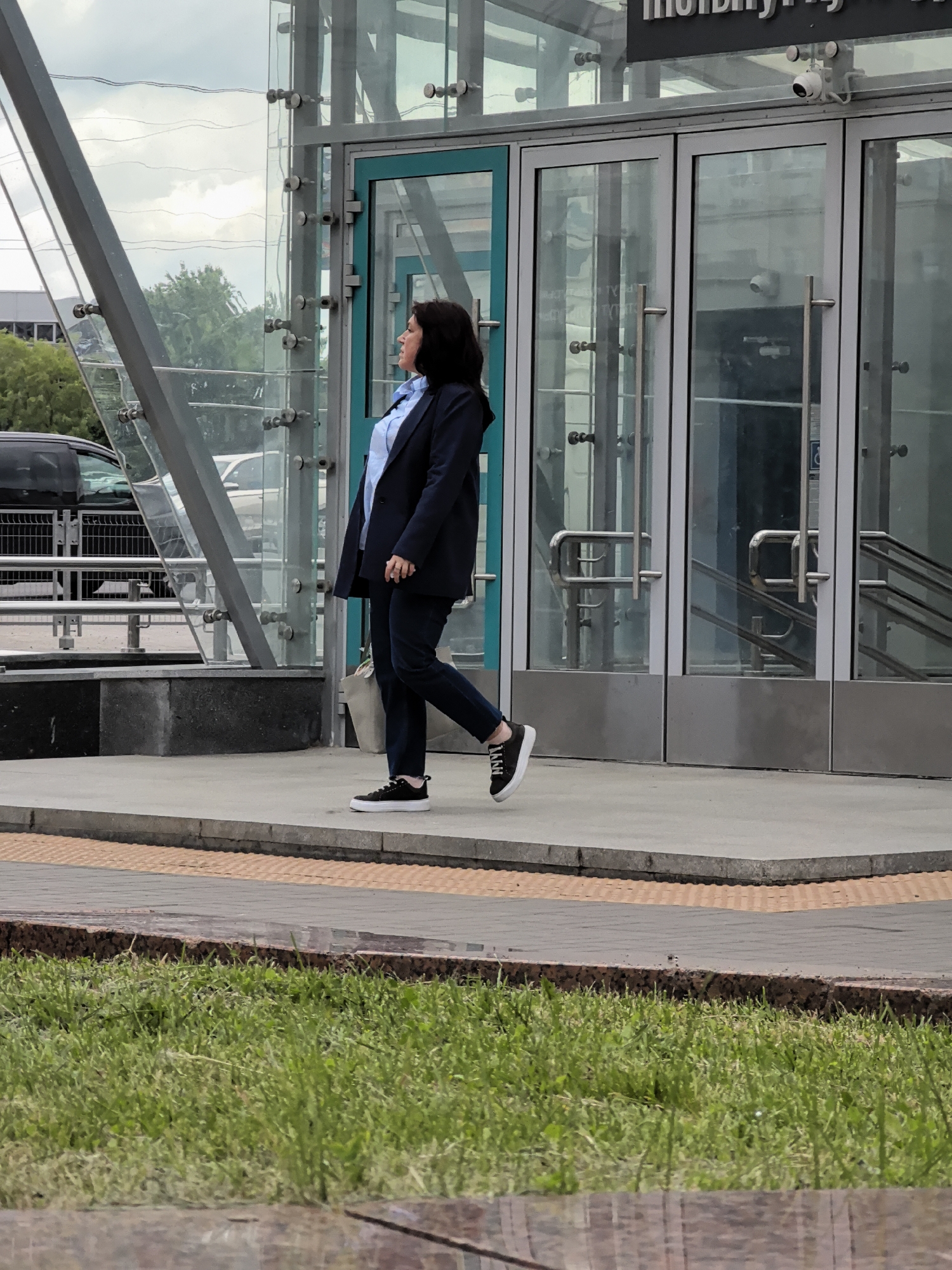 A medium-shot, eye-level, full-body outdoor photograph shows a woman walking on a paved walkway in front of a modern building with glass walls and doors. The woman, with dark hair and fair skin, is wearing a dark blue blazer over a light blue collared shirt and dark blue pants. She has on black sneakers with white soles and laces. Her left hand is slightly extended, and she is looking to her left. A light-colored tote bag is slung over her left shoulder. The building appears to be an entrance to a public space, with metal railings and a ramp visible through the glass. Security cameras are mounted above the entrance. The walkway has a yellow tactile paving strip along its edge. In the foreground, out of focus, is a patch of green grass. The background shows trees and what appears to be a cloudy sky. The scene is captured in daylight.
