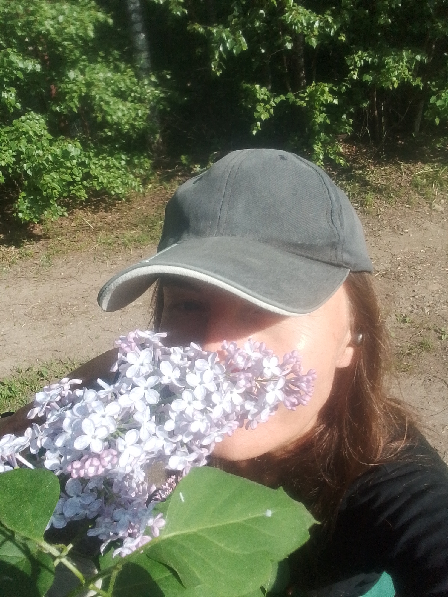 A person wearing a gray baseball cap holds a large bunch of light purple and white lilac flowers close to their face. The person is wearing a black long-sleeved shirt. The location is a dirt path with sparse vegetation. The background shows dense green foliage and trees, likely in the vicinity of Снопок Новый, Russia.