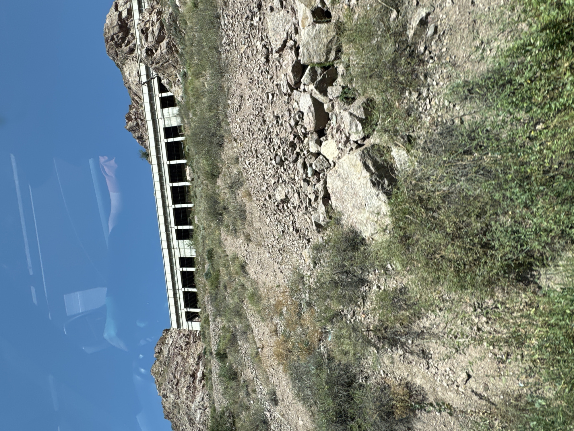 The image shows a highway overpass built into a rocky hillside in Cholok, Kyrgyzstan. The overpass is a concrete structure with multiple arched openings, supported by pillars. The hillside is covered in loose rocks, gravel, and sparse vegetation, including bushes and dry grasses. The sky is clear and blue, with reflections of what appears to be a vehicle's interior visible on the surface, suggesting the photo was taken from inside a car or bus. There are no people visible in the image.