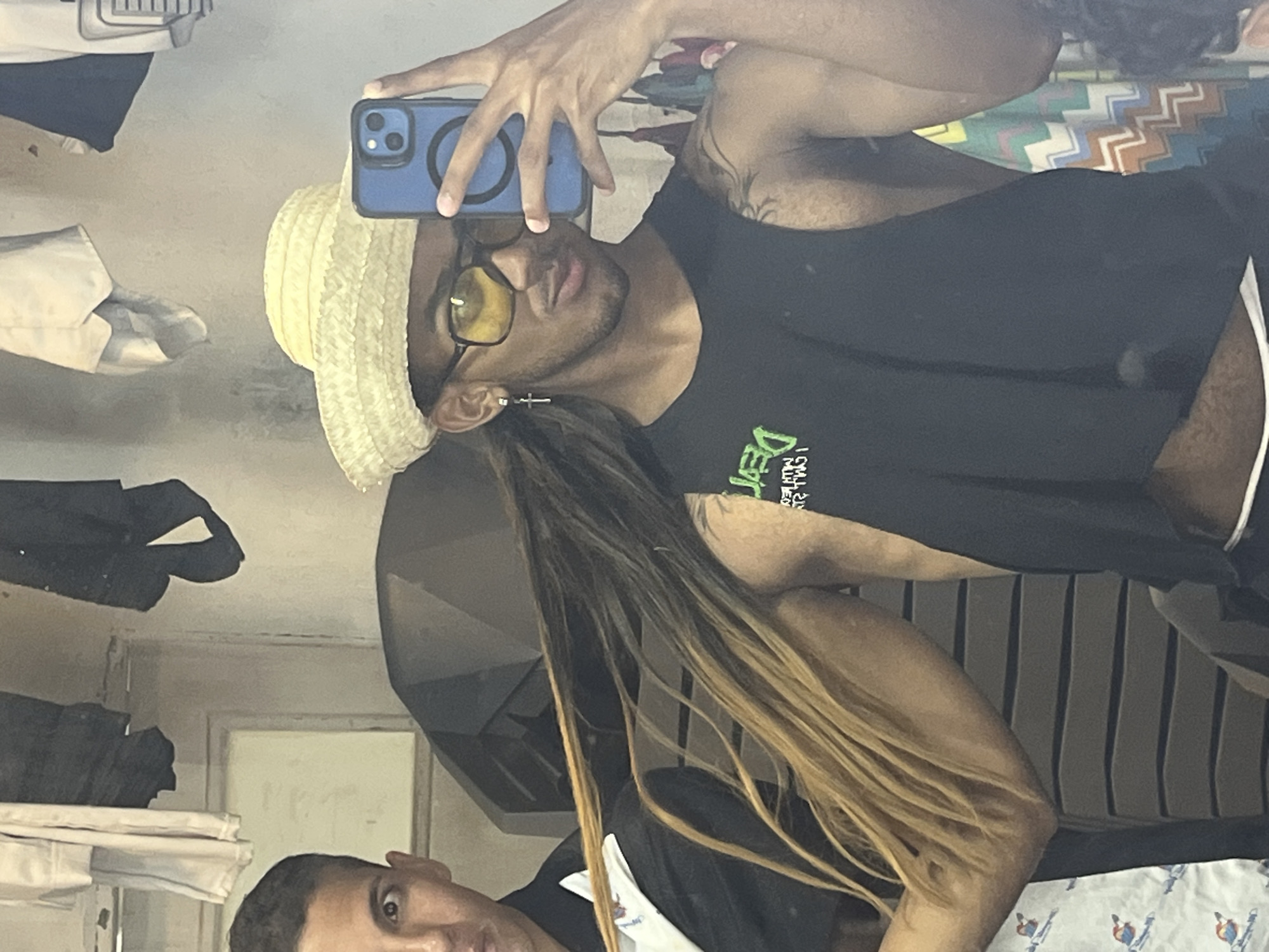 The image shows a selfie taken in front of a mirror. A dark-skinned person, likely male, is wearing a straw hat, dark sunglasses, and a black sleeveless shirt. He is holding a smartphone with a blue case. Reflected in the mirror is another person with long, highlighted hair. In the background, there are shelves with folded clothes, a window, and patterned bedding. There is no information to confirm or deny that the location is in the city of Porlamar, Venezuela.