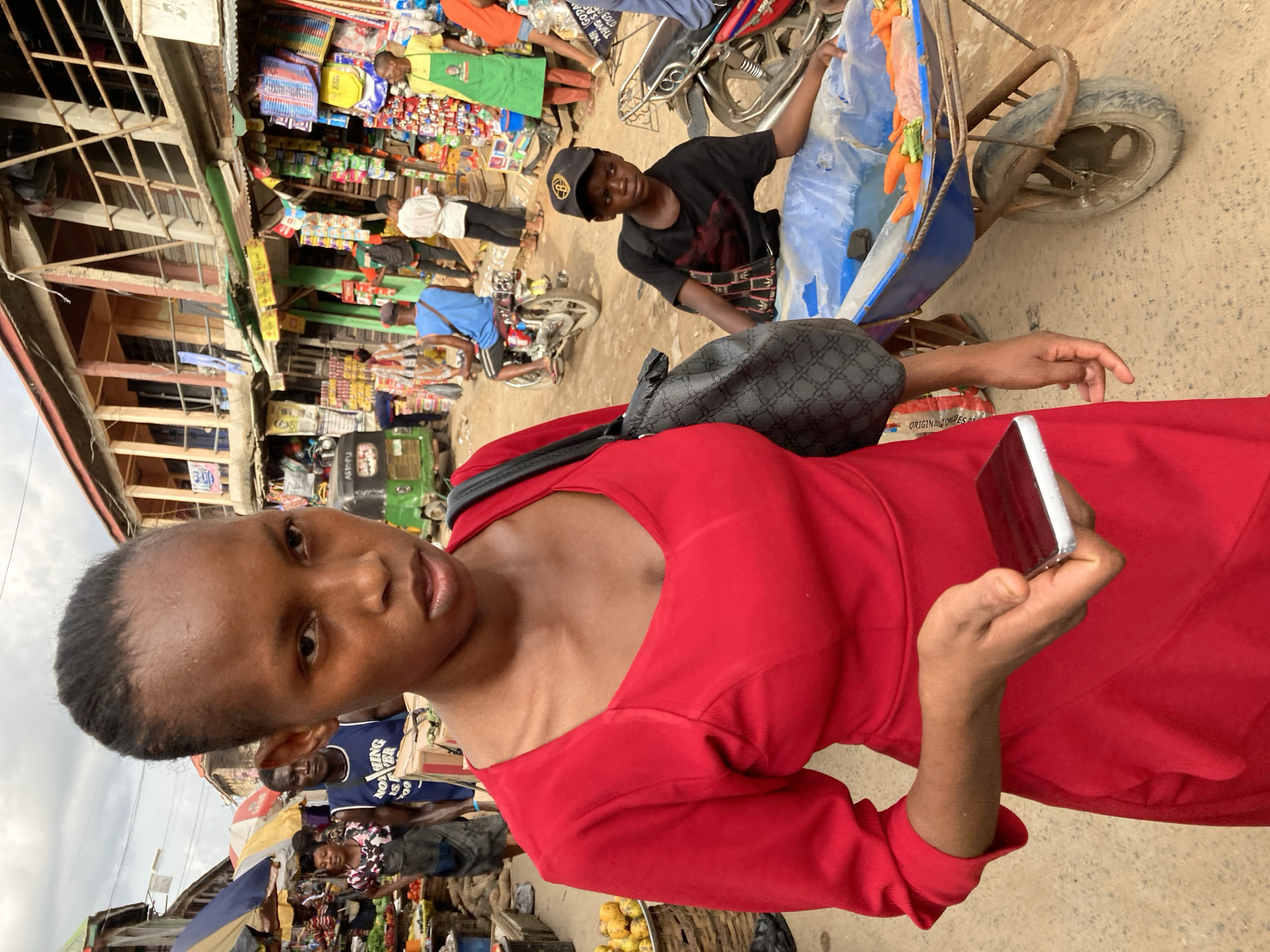 The image depicts a woman in a red dress holding a mobile phone, seemingly taking a photograph in a bustling market environment. The background reveals market stalls with various goods on display, including produce and packaged items. A young man wearing a black shirt and a cap is standing next to a blue wheelbarrow containing carrots. There are other individuals milling about in the market, suggesting a lively atmosphere. Based on the prompt, the location is Ura Nta Umuarandu, Nigeria.