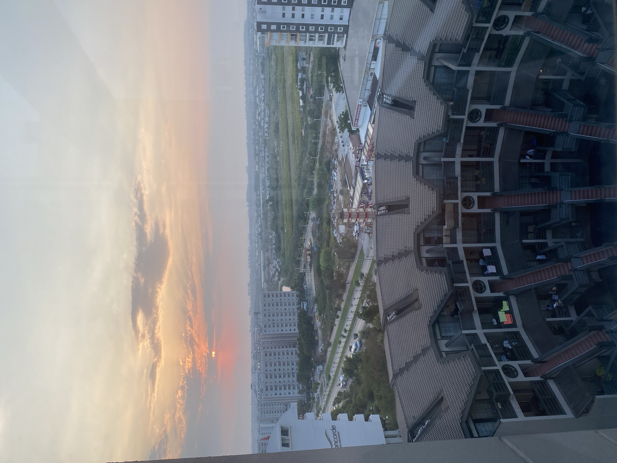 This is a photograph of the city of Aksu, Turkey, at sunset. The foreground of the image is dominated by the curved facade of a multi-story building with a tiled roof. This building features external staircases and balconies, with laundry visible hanging on some of the balconies.
Beyond this building, a road runs horizontally across the scene, with several vehicles, including white vans and cars, traveling on it. To the left of the road, there is another building with the name "concorde" and "DE LUXE RESORT" visible on its side. Further into the background, a series of large, multi-story apartment buildings stretches across the cityscape. Beyond these buildings, the landscape transitions to a rural area with green fields and scattered houses. The horizon is marked by a hazy skyline of distant buildings and hills.
The sky is a prominent feature, filled with soft, warm colors of sunset. The sun, partially obscured by clouds, casts a warm orange glow, with rays of light breaking through. The overall impression is of a residential area at dusk.