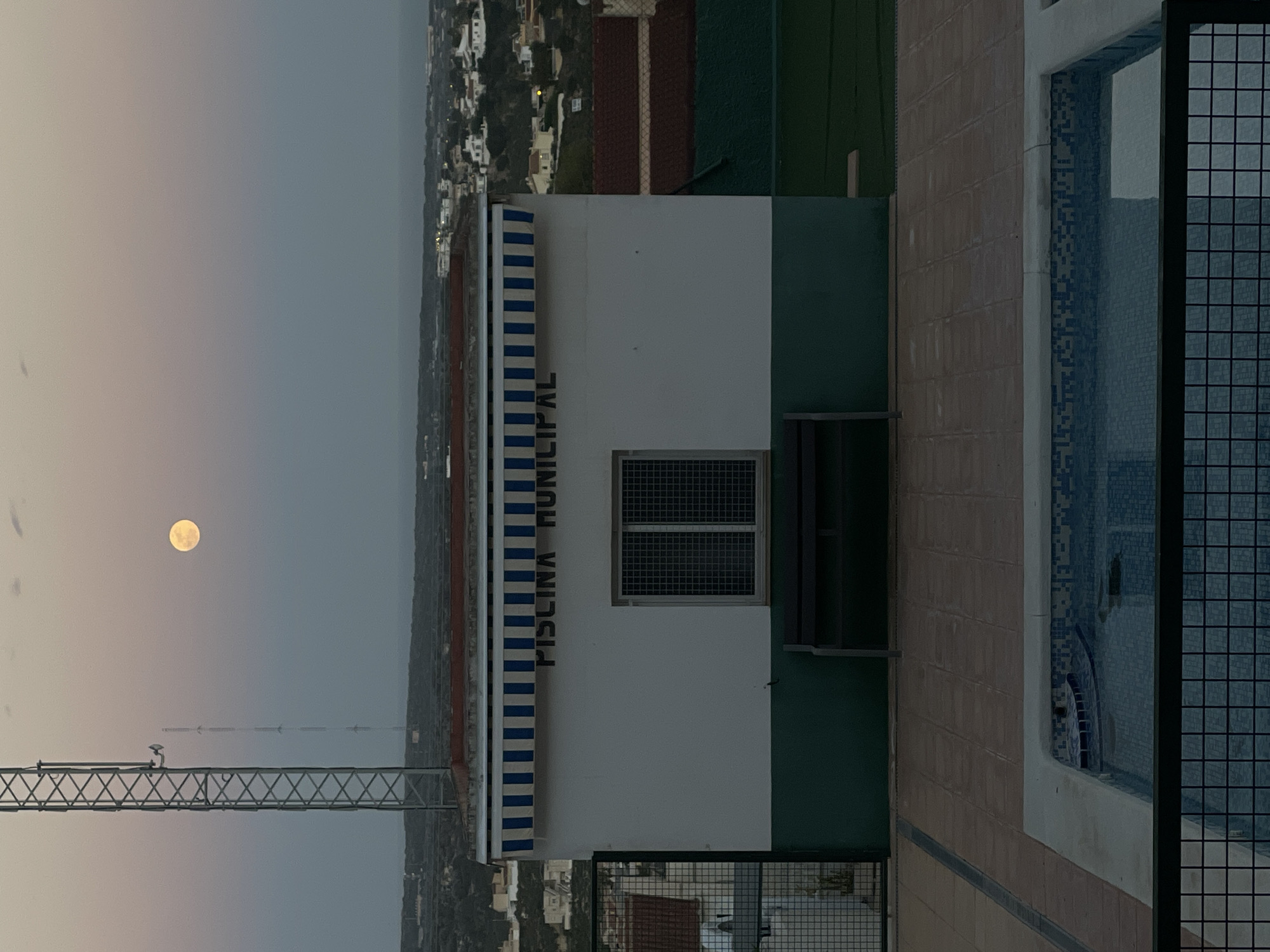 The image depicts a building in Rojales, Spain, specifically the "Piscina Municipal," which translates to Municipal Pool. The building is painted white and green with a blue and white striped awning. A window with a grid pattern is visible on the building. The image includes part of a pool area, enclosed by a metal grid. A full moon is visible in the sky. A communications tower is also visible in the image.