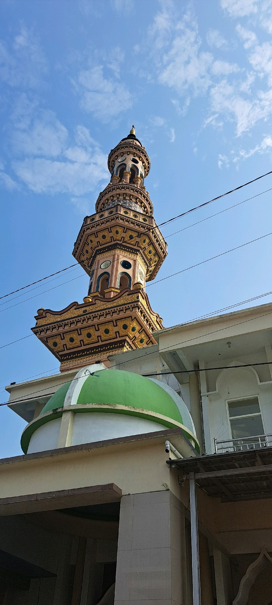 A minaret of a mosque towers over a green dome against a bright blue sky with scattered clouds. The minaret is ornate with multiple levels and decorative patterns in gold, pink, and blue. Below it, a portion of the mosque building is visible, including a white wall with a window and a balcony. Power lines stretch across the sky in front of the minaret. The image appears to be taken from a low angle, emphasizing the height of the minaret. The location is the city of Duyung, Indonesia.