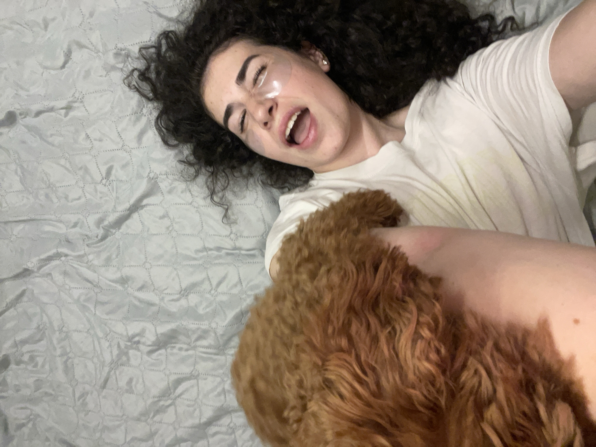 In a bedroom in Sofia, Bulgaria, a woman with dark, curly hair lies on a light blue, quilted bedspread. She is wearing a white t-shirt and has under-eye patches on. Her mouth is open in a scream or laugh, and her eyes are closed. A small, fluffy brown dog, possibly a poodle mix, rests its head on her chest. The lighting suggests it is daytime, but the exact time and source of light are not discernible.