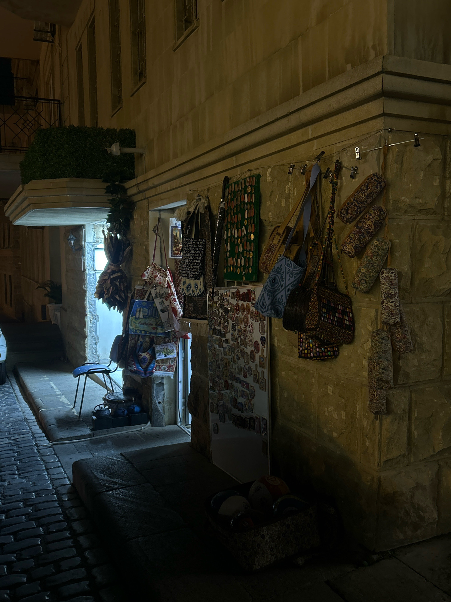 In a narrow cobblestone alleyway in Sabail Raion, Azerbaijan, a small shop is displayed outdoors. Various handcrafted items, including bags and textiles adorned with intricate patterns, are hung on the stone wall of the building and in the doorway. A blue stool stands near the entrance, and a collection of smaller decorative items is displayed on a white board. The scene is illuminated by artificial light, creating shadows and highlighting the textures of the stone and merchandise. The shop appears to be closed, with no people visible in the immediate vicinity. The architecture suggests an older part of the city, with stone buildings and a traditional alleyway design.