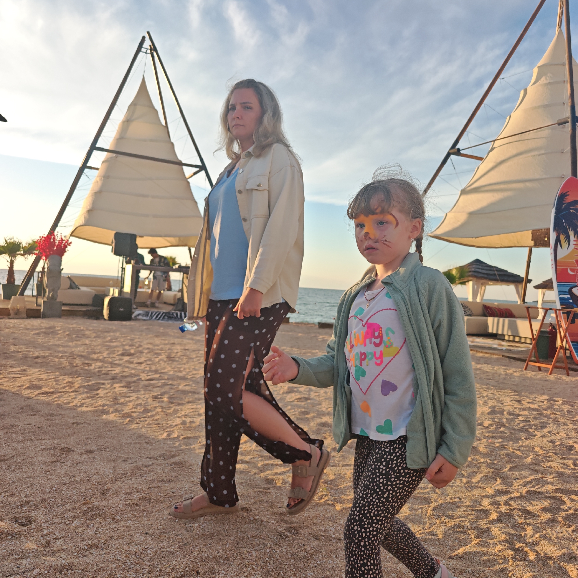 The image shows a woman and a young girl walking on a sandy beach, likely in the city of Shchelkino, Ukraine, given the information provided. The woman, on the left, has blonde hair and is wearing a light-colored jacket over a blue top and brown polka-dotted pants. The girl, on the right, has her hair in braids and sports face paint resembling a cat or lion. She is wearing a green cardigan over a white t-shirt with colorful hearts and patterned leggings. In the background, there are two large, sail-like structures, possibly tents or awnings, set up on the beach. A body of water, likely the sea, is visible beyond the beach. Further in the background, a DJ setup with speakers and a person appears to be present under one of the awnings, suggesting some sort of event or gathering. The overall lighting suggests it is late afternoon or early evening.