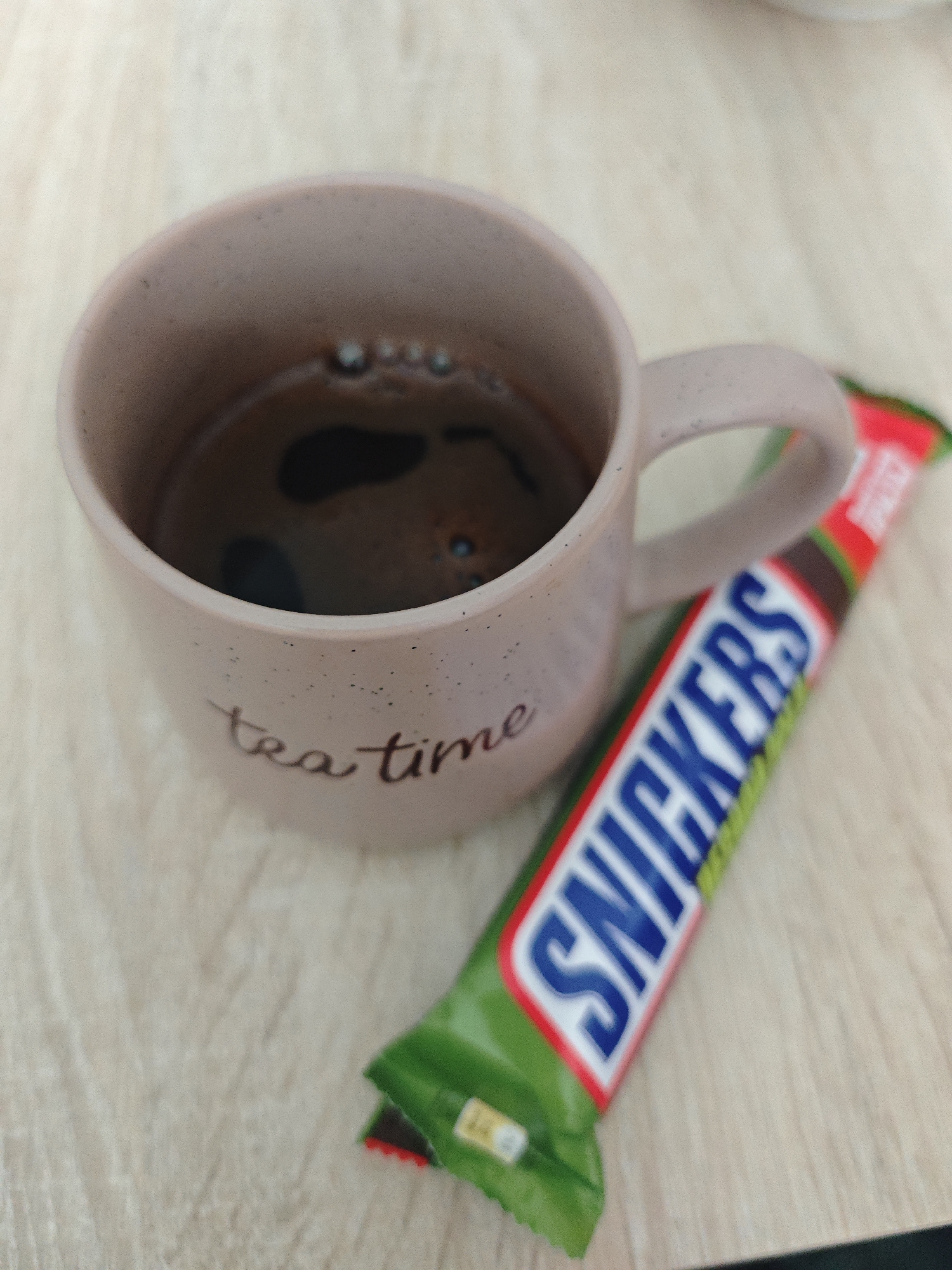 A close-up shot shows a pink mug with "tea time" written on it, filled with dark liquid, and a Snickers candy bar. The mug and candy bar are positioned on a light-colored, textured surface, possibly a table. The background is blurred and out of focus. There are no people or other significant objects visible in the frame. The lighting appears to be natural and overhead, casting soft shadows.