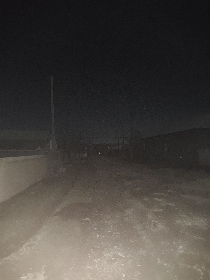 A dimly lit rural scene at night. A dirt road stretches into the distance, bordered by unseen fields and fences. A tall, slender pole stands to the left. Buildings are faintly visible on either side of the road, shrouded in darkness, suggesting a small village. The sky is an inky black, blending seamlessly with the landscape. The ground appears uneven, scattered with rocks or debris. The overall atmosphere is one of isolation and quiet solitude.