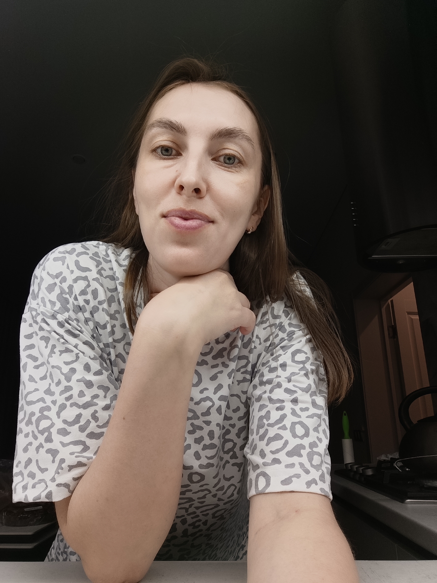 A woman with fair skin and brown hair, wearing a white and gray animal-print shirt, rests her chin on her hand. She is indoors, appearing to be in a kitchen setting. In the background is a stove with a kettle on it, and a kitchen ventilation hood is visible above. The woman is located in Chaikovsky, Russia.