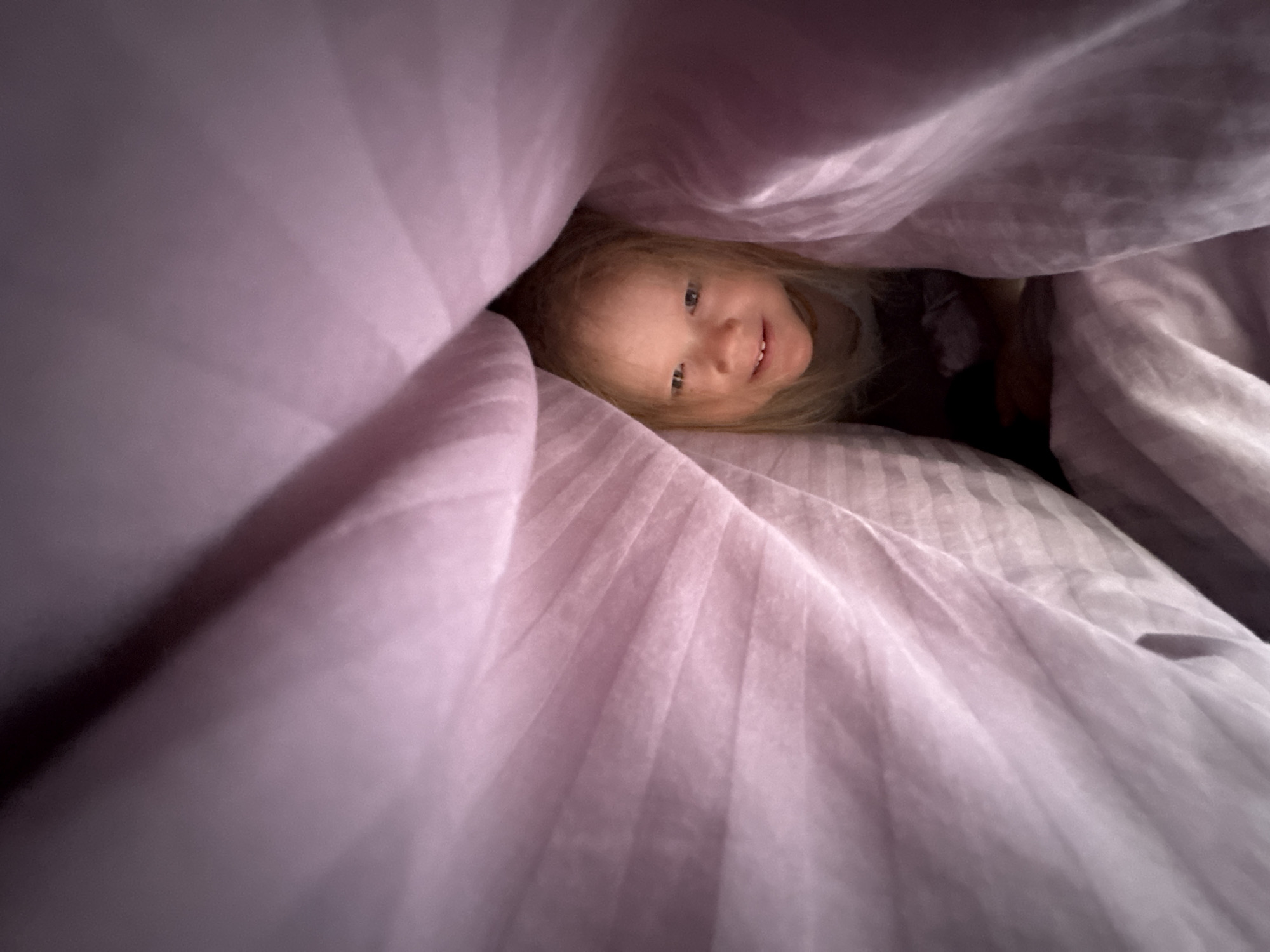 A young child with blonde hair is partially visible, peeking out from under purple striped bedding. The child is smiling slightly. The bedding dominates the frame, obscuring most of the surrounding environment. The location is unknown.