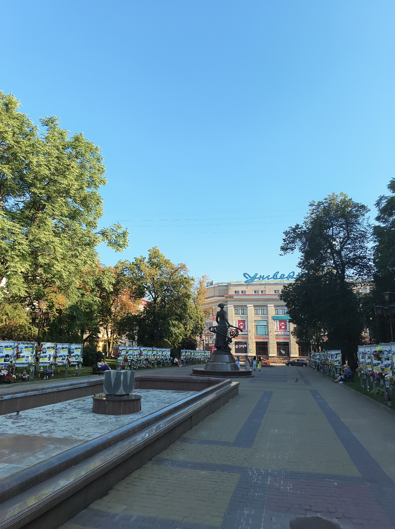 The image captures a wide-angle view of a public square in Ternopil, Ukraine, likely on a sunny day given the bright sky. In the foreground, a wet fountain with a sculptural element shaped like stylized tulips is prominent. The fountain is surrounded by a stone embankment, and adjacent to it is a paved walkway.
To the left of the fountain, a line of posters is displayed, each featuring portraits and names, likely memorializing fallen soldiers. These posters are adorned with flowers. Further to the left, a large, leafy tree provides shade.
In the background, a multi-story building with "Універмаг" (Department Store) in large blue lettering on its facade stands. Several storefronts are visible on the ground floor, including "ZARA" and "Jack&Jones". A statue of a woman is positioned in front of the building, near the memorial posters.
Several people are present in the scene, some sitting on benches near the posters and others walking along the paved path. The environment suggests a somber yet public space.