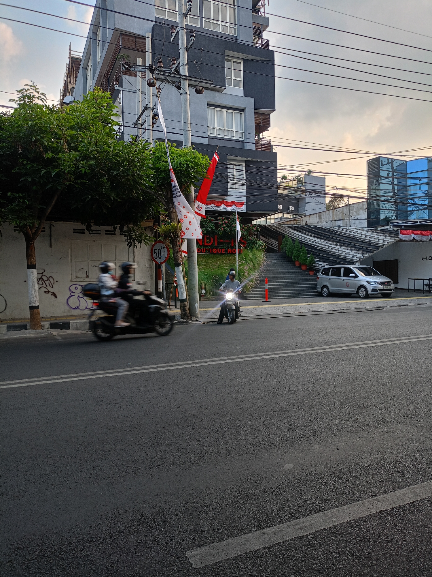 In the city of Batu, Indonesia, a street scene unfolds with a modern building as a backdrop. The building features a combination of gray and dark-colored facades with several floors, adorned with numerous windows. In front of the building, a set of wide, tiered stairs leads up to an entrance. To the right of the stairs, a silver minivan is parked.
A paved road occupies the foreground, with two white lines marking the lanes. On the road, two motorcycles are visible. One motorcycle, closer to the left side of the image, is carrying two people, both wearing helmets. This motorcycle is blurred due to motion. To the right, a single rider on a motorcycle, also wearing a helmet, is traveling in the same direction. This motorcycle's headlight is illuminated.
Alongside the road, a sidewalk is present, with a speed limit sign indicating "50 km/h." Several trees with lush green foliage are planted along the sidewalk, and a utility pole with numerous wires stretches across the scene. Flags, one white with red accents and another partially visible red and white flag, are also displayed near the utility pole. Graffiti is visible on a wall to the left of the road. In the distance, another building with blue glass windows can be seen. The sky above is overcast, suggesting a cloudy day.