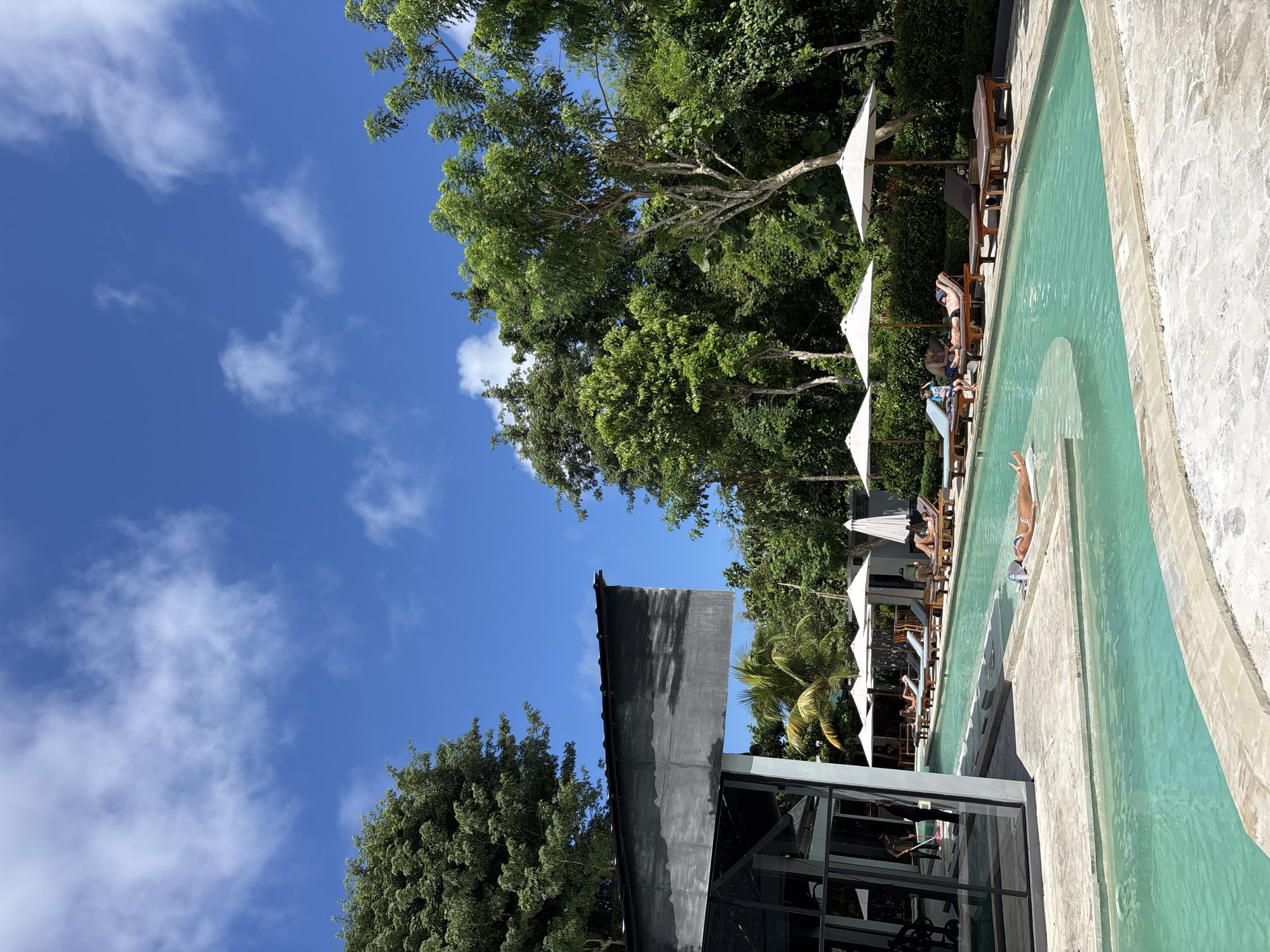 The image shows a swimming pool scene in Kuta Selatan, Indonesia. A person is diving into the light blue pool. Several white umbrellas and lounge chairs are positioned along the pool's edge where people are relaxing. The pool is next to a building with large windows. Green trees and a clear blue sky with scattered clouds form the background.