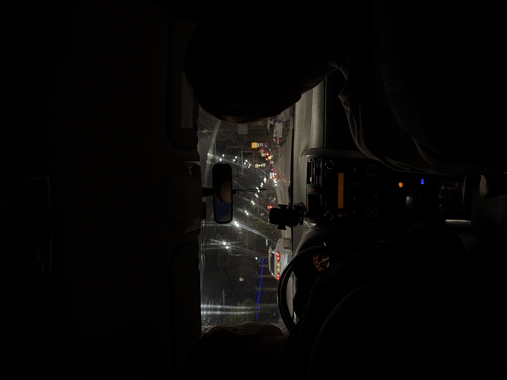 A nighttime scene from inside a vehicle, likely a car, shows a driver looking out the front windshield at a city street with traffic lights. The view through the windshield reveals a wet road surface reflecting the lights, suggesting recent or ongoing rain. The road is lined with streetlights and what appear to be buildings or structures beyond the immediate roadway. The interior of the car is dimly lit, showcasing the dashboard and steering wheel. The overall image is dark and the specific location in Izhevsk, Russia cannot be determined.