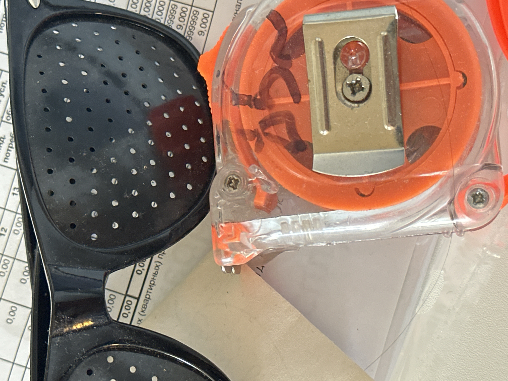 The image shows a close-up of a pair of black pinhole glasses, a portion of a roll of measuring tape, and some paper documents. The glasses are positioned over the documents. The measuring tape is orange and clear plastic, and it has some black markings on it, possibly a signature or initials. The paper documents appear to be a form or a report with columns of numbers and text. The background is out of focus, but it seems to be a flat surface, possibly a desk or table. There are no people visible in the image. The image does not provide any information about the city of Nevinnomyssk, Russia.
