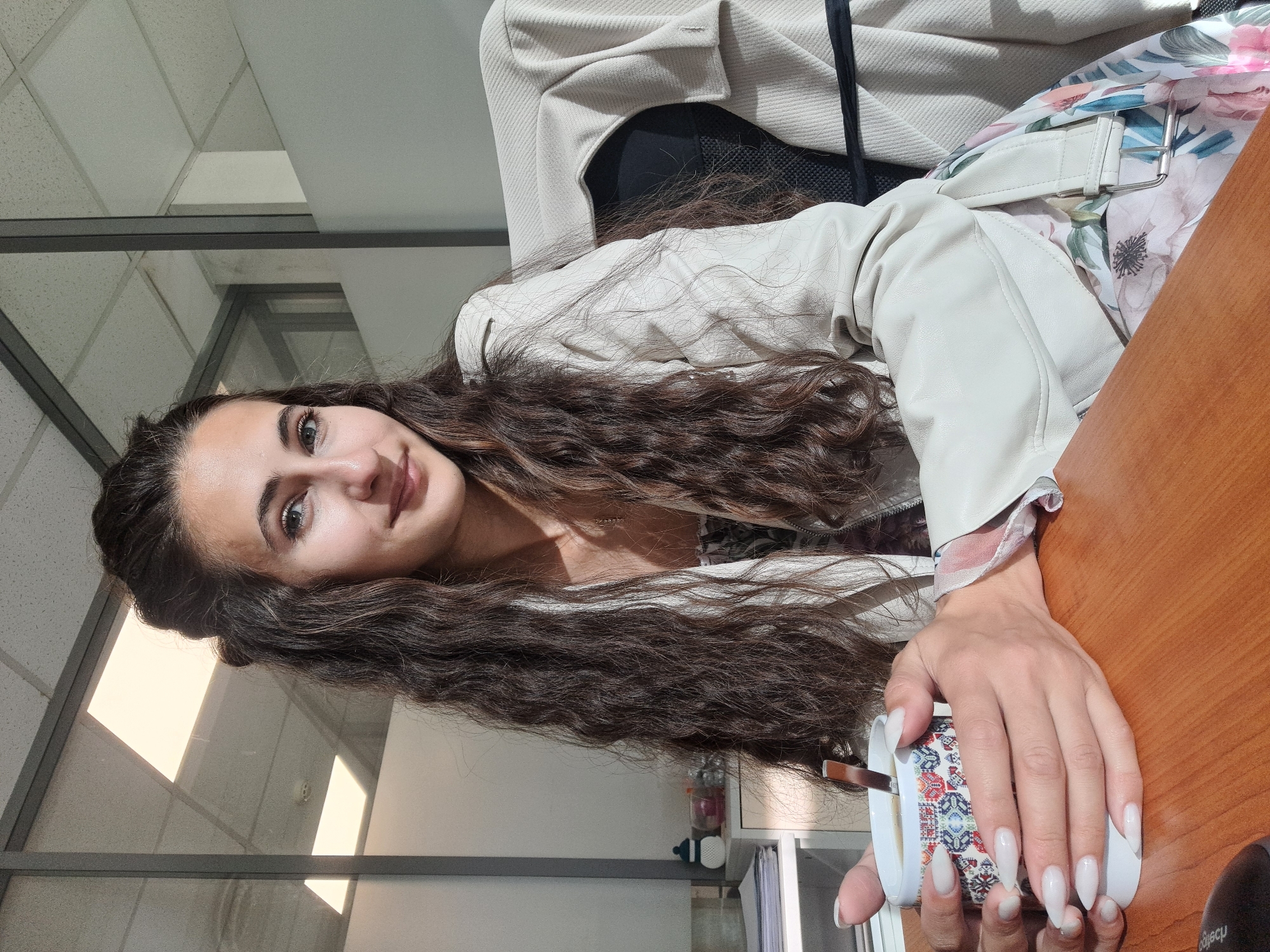 A woman with long, dark, curly hair sits at a wooden desk in an office. She is wearing a light-colored jacket over a floral-patterned dress. She holds a white mug with a colorful geometric pattern, containing a beverage and a spoon. Her fingernails are long and painted white. The background shows office partitions with frosted glass and fluorescent lighting, typical of a modern office space. The sunlight streaming in creates shadows and highlights on the woman and her surroundings. The scene is likely set in an office in Sofia, Bulgaria, given the contextual information.