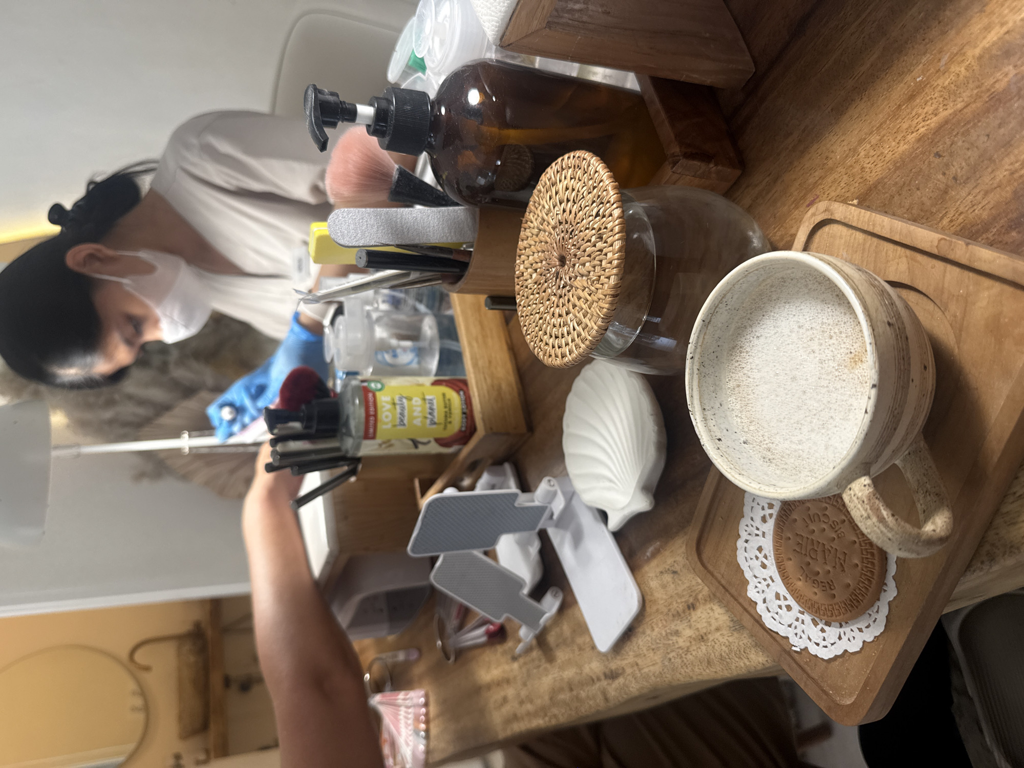 On a wooden surface, a white ceramic mug rests on a small wooden tray over a white lace doily and a cookie labeled "REGAL MARIE BISCUIT." Adjacent to the mug is a white seashell and a glass container with a woven lid. A wooden organizer holds makeup brushes, nail files, and an amber-colored pump bottle. An "Love beauty and planet" branded bottle is visible near an assortment of scattered makeup accessories. A person wearing a mask and blue gloves is seen in the background. The location is unidentified.