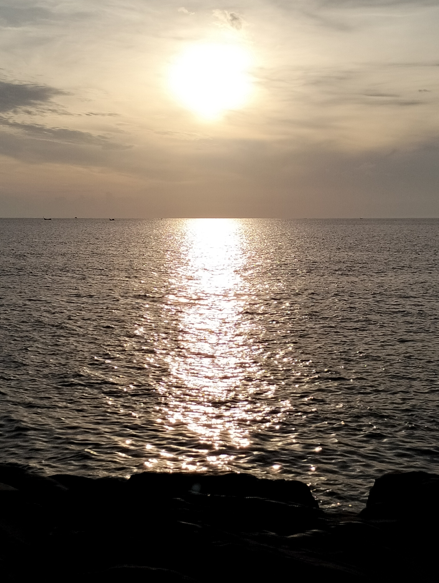 The image shows a view of the sea at sunset. The sun is low in the sky, casting a bright reflection on the water. There are several small boats visible on the horizon. The foreground is in silhouette, with dark rocks and land. The location is the shore of Lhokseumawe, Indonesia, looking out over the sea.