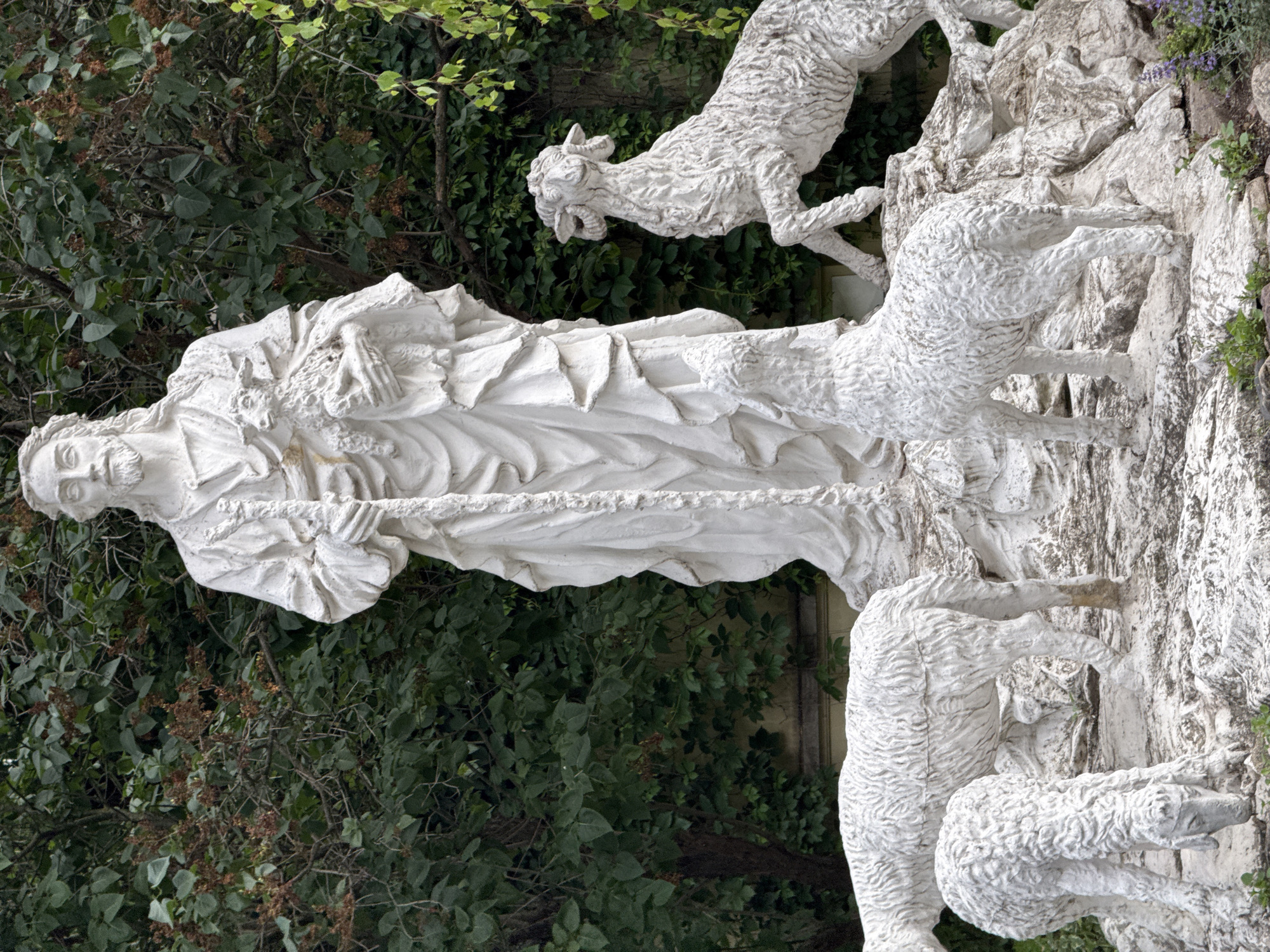 This is a full outdoor shot of a white stone sculpture of Jesus Christ holding a lamb, standing amidst several other lamb sculptures. The sculpture is located in the Alexander Garden in Moscow, Russia. Jesus is depicted with a long beard and flowing robes, holding a staff in his left hand and a lamb in his right arm. The lambs are in various poses: one is held by Jesus, another is standing in front of him, and two more are in the foreground, one grazing and the other standing. The sculpture is set against a backdrop of lush green trees and foliage. The overall impression is one of peace and serenity.
