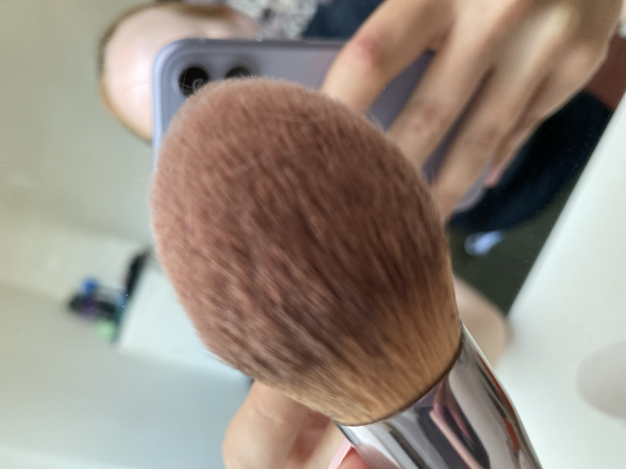 The image shows a close-up of a makeup brush, with its fluffy, brown bristles in sharp focus. The handle of the brush appears to be made of silver-toned metal. In the blurred background, a reflection in a mirror reveals a person holding a purple smartphone and their head. The person has fair skin and light-colored hair. The environment appears to be indoors, possibly a room with a white surface in the foreground and some indistinct objects in the background. There is no information within the image to suggest a location in Kisliakovskaya, Russia.
