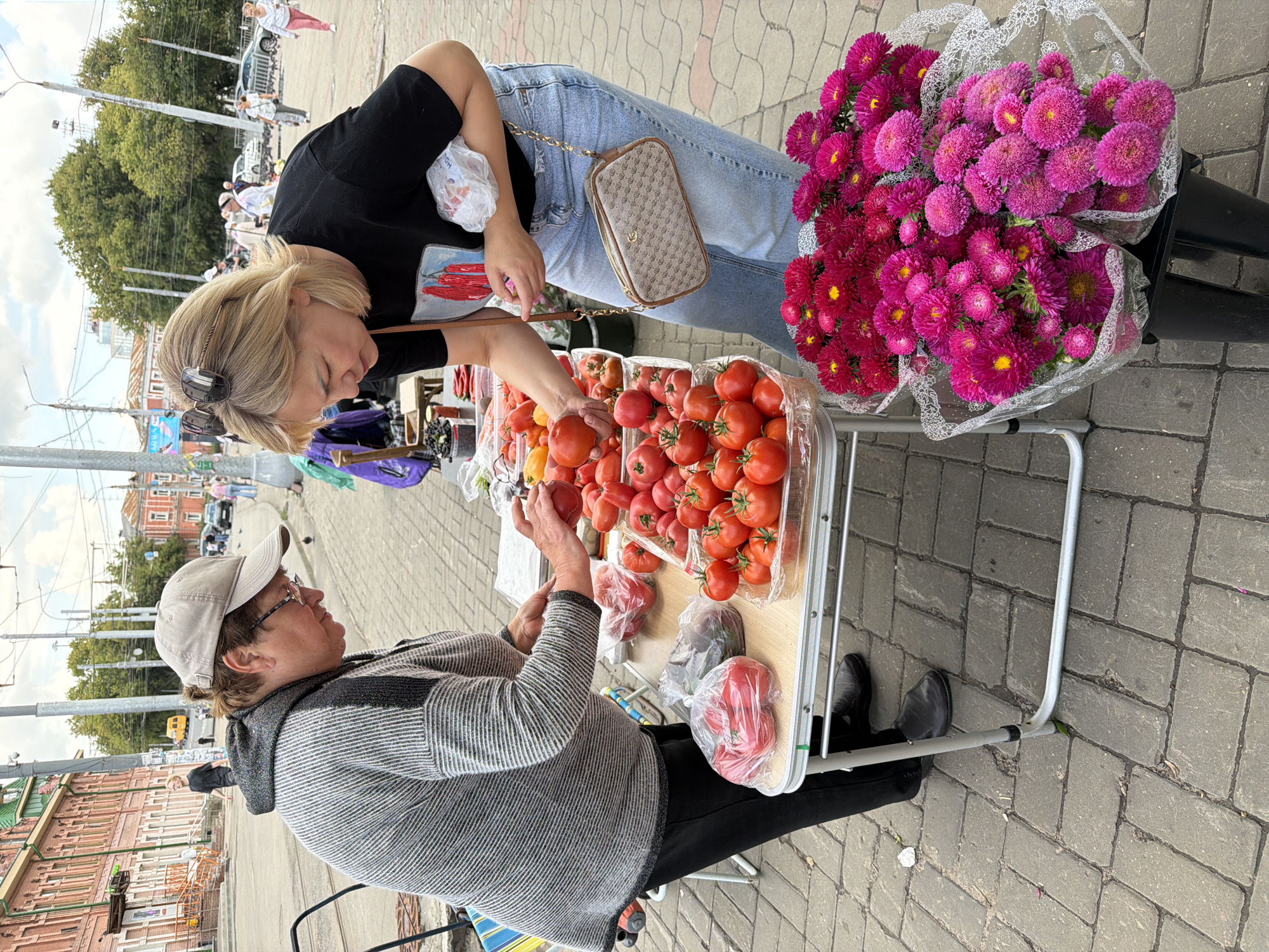 In Nizhny Novgorod, Russia, a vendor wearing a grey striped sweater and a tan baseball cap stands behind a table laden with ripe tomatoes. A customer, a woman with blonde hair, wearing a black t-shirt and blue jeans, selects a tomato from the display. She holds a plastic bag in her left arm and a small patterned purse with a chain strap hangs from her shoulder. The table also holds plastic bags of tomatoes, and in the foreground, bunches of vibrant pink and red chrysanthemums are displayed in clear plastic. The scene takes place outdoors on a paved walkway, likely a street market, with buildings and trees visible in the background, suggesting an urban setting.