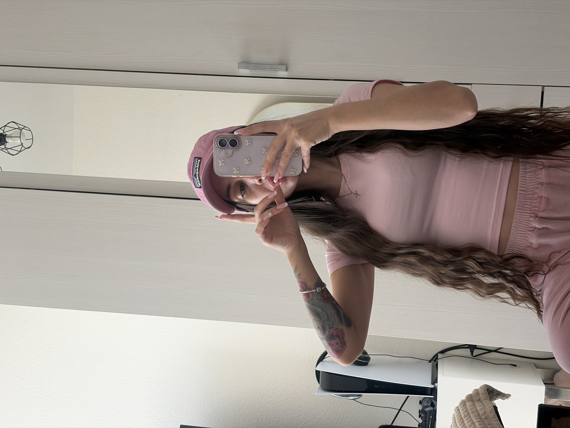 A woman in a pink outfit and cap is taking a selfie in front of a mirror in an indoor setting. The room features light-colored walls, a white wardrobe with silver handles, and a shelf where a Playstation 5 is placed. The woman is holding a phone with a light-colored case with floral patterns. She has long wavy brown hair and a tattoo on her left arm. The reflection in the mirror shows additional details of the room, including a geometric wireframe decoration. The location cannot be identified as Saint Petersburg, Russia.