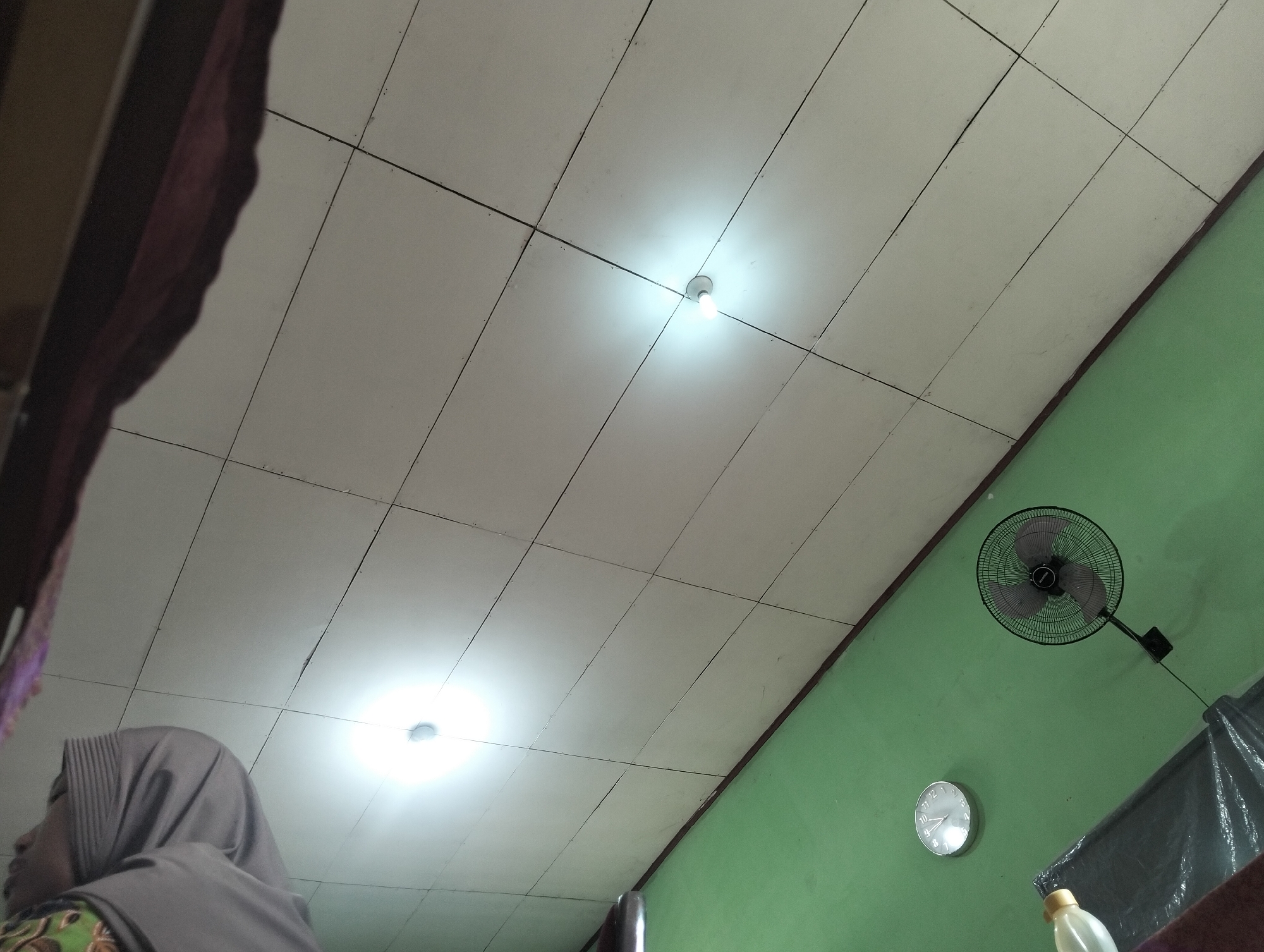 The image depicts an indoor scene, likely a room, with a focus on the ceiling and a portion of a green-painted wall. Two ceiling lights are visible, one brighter than the other, illuminating the white, tiled ceiling. On the green wall, a black oscillating fan is mounted, and a silver analog clock is also present. In the lower left of the frame, the head and shoulder of a person wearing a grey hijab and a patterned garment are visible, looking towards the right. The overall lighting suggests it is daytime, but the artificial lights on the ceiling are also active. The background shows the corner of a room where the ceiling meets the wall. A dark curtain or covering is draped on the far right of the wall, partially obscuring another object that might be a bottle. The presence of common household items like a fan and clock, along with the visible person, suggests a domestic setting. The location is identified as Banjar Agung, Indonesia.