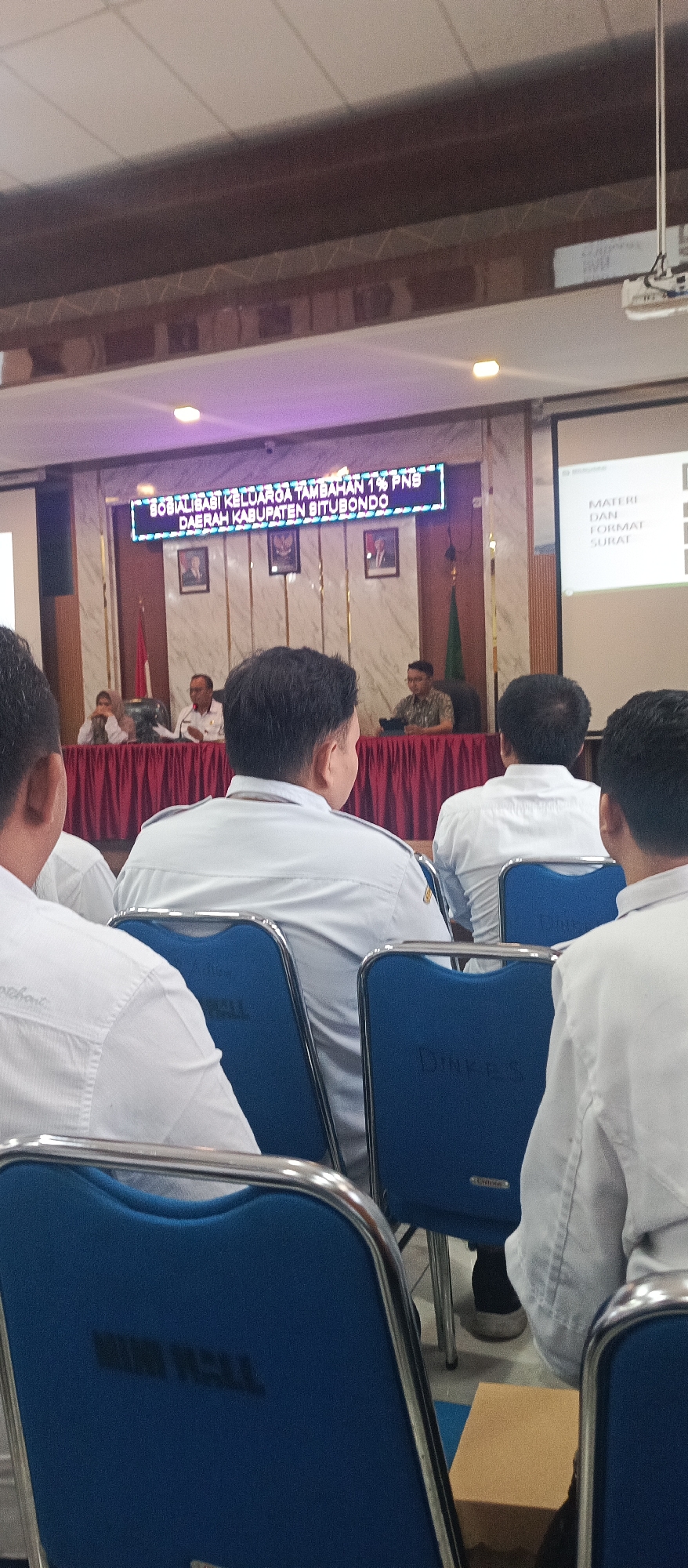 The image shows an indoor gathering, likely a meeting or seminar, taking place in a room that appears to be a government or public office in Situbondo, Indonesia. In the foreground, several individuals are seated, viewed from behind, wearing light-colored shirts, some with a semi-formal or uniform appearance. The chairs they occupy are blue with metal frames and have text visible on them, possibly identifying the organization or event.
In the mid-ground, a table is set up with several individuals seated behind it, facing the audience. A prominent digital display board above the table shows text in Indonesian, which reads "SOSIALISASI KELUARGA TAMBAHAN 1% PNS DAERAH KABUPATEN SITUBONDO". This translates to "Socialization of Additional 1% PNS Family in Situbondo Regency". PNS is likely an abbreviation for Pegawai Negeri Sipil, meaning civil servant. The backdrop behind the table features framed portraits and a green flag.
To the right of the display board, a projector screen is visible, displaying text that appears to be related to "MATERI DAN FORMAT SURAT" (Materials and Letter Format). The environment is lit by overhead lighting, with some purple lighting emanating from the digital display. The overall scene suggests an official event or training session related to civil servants in Situbondo Regency.