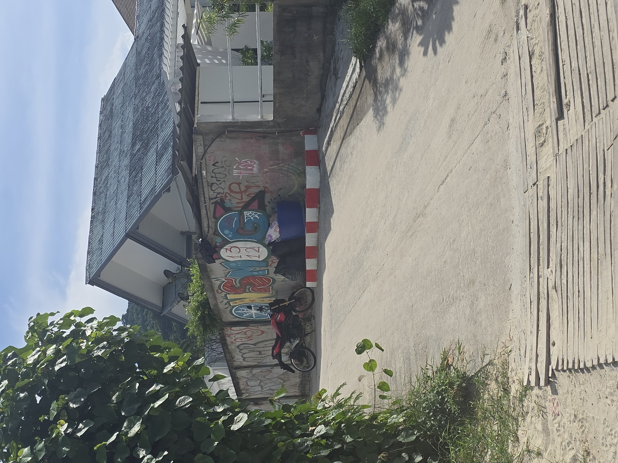 A dirt road slopes upwards in front of a concrete wall covered in graffiti. To the left, a motorcycle is parked. To the right, black trash bags are piled near a blue plastic bin. Beyond the wall is a building with a corrugated metal roof and white siding. Lush green foliage obscures the top left corner of the frame. In the distance, a hillside covered in trees is visible under a blue sky with scattered clouds. A wooden walkway made of planks is laid across the road in the foreground. The scene is located in Baan Chaweng Noi, Thailand.