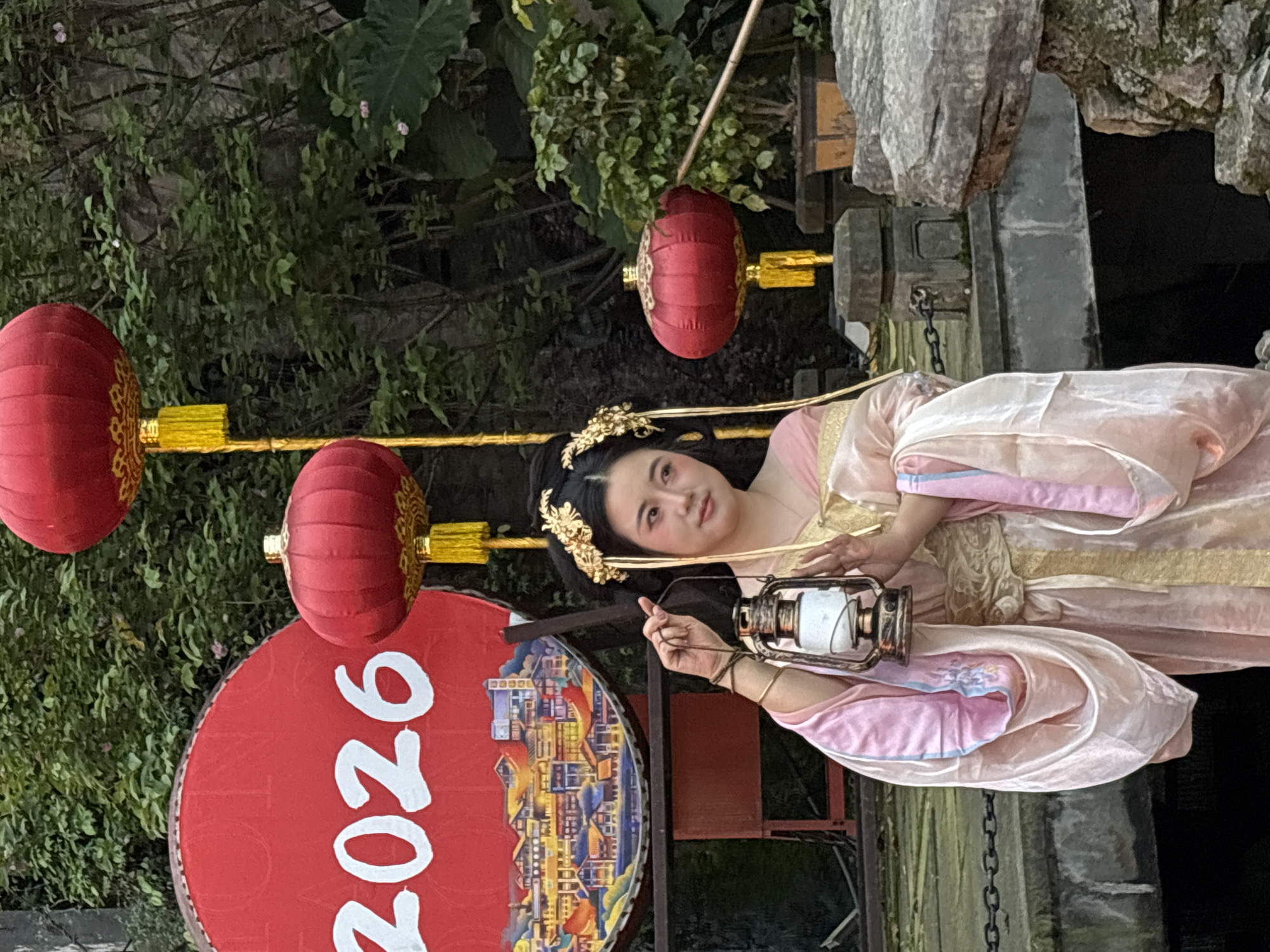 A person dressed in traditional Chinese clothing stands outdoors in Yuzhong District, China. The person is holding a lantern. Red lanterns hang nearby, and a sign with "2026" is visible. Trees and vegetation are in the background. The person appears to be near a bridge or stone structure, with water below.