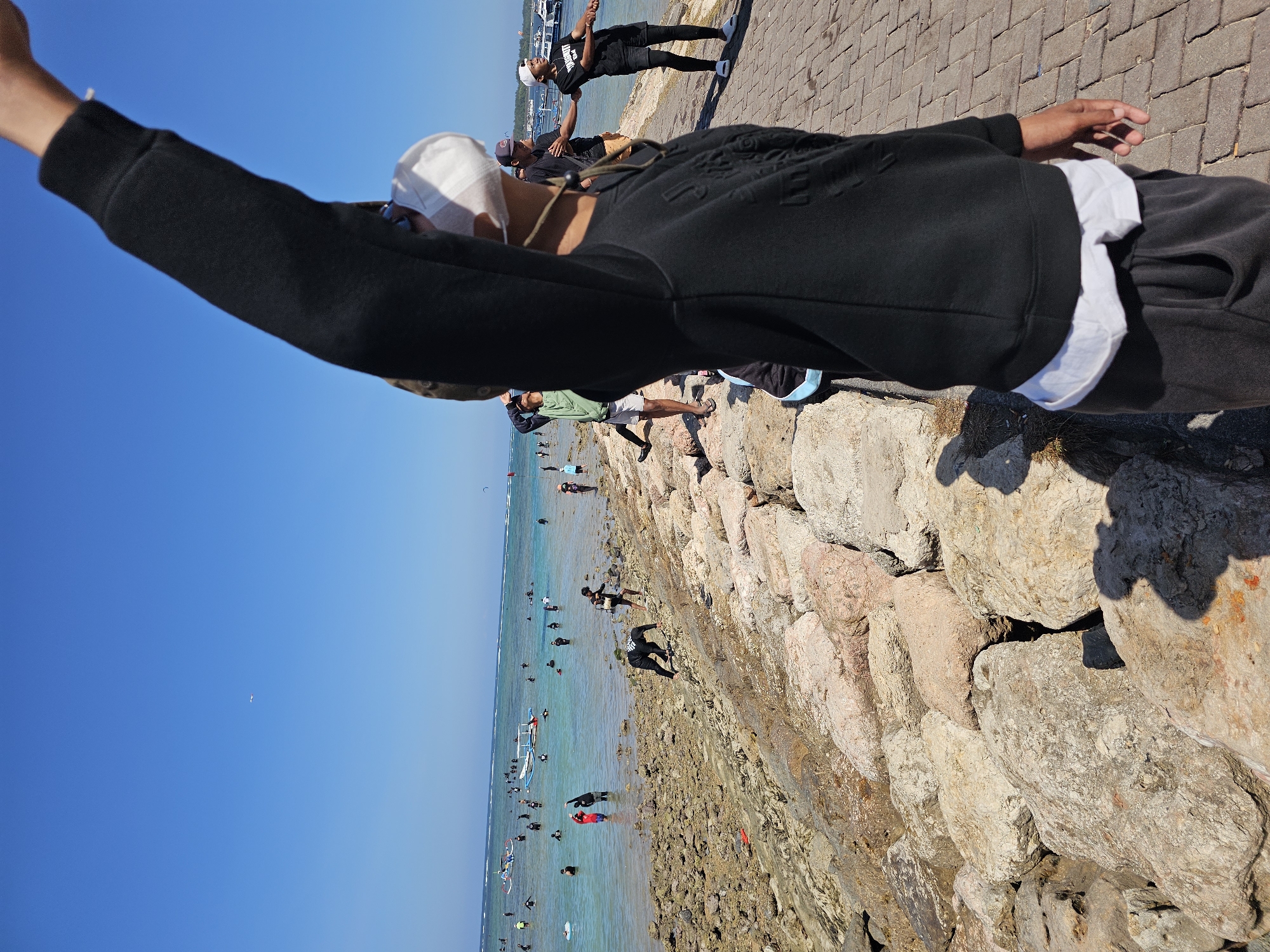 The image captures a sunny day at a beach in Denpasar, Indonesia. In the foreground, a person is seen from behind, wearing a black sweatshirt, a white face mask, and dark pants. Their right arm is raised, reaching towards the sky. The person is standing on a paved walkway. To the left of the walkway is a rocky shore, extending into the shallow, turquoise water. Several people are visible in the water, some swimming and others standing. A few boats are also present in the sea. The sky is clear and blue with a distant airplane. The scene depicts a lively beach environment with people enjoying the water and shore.