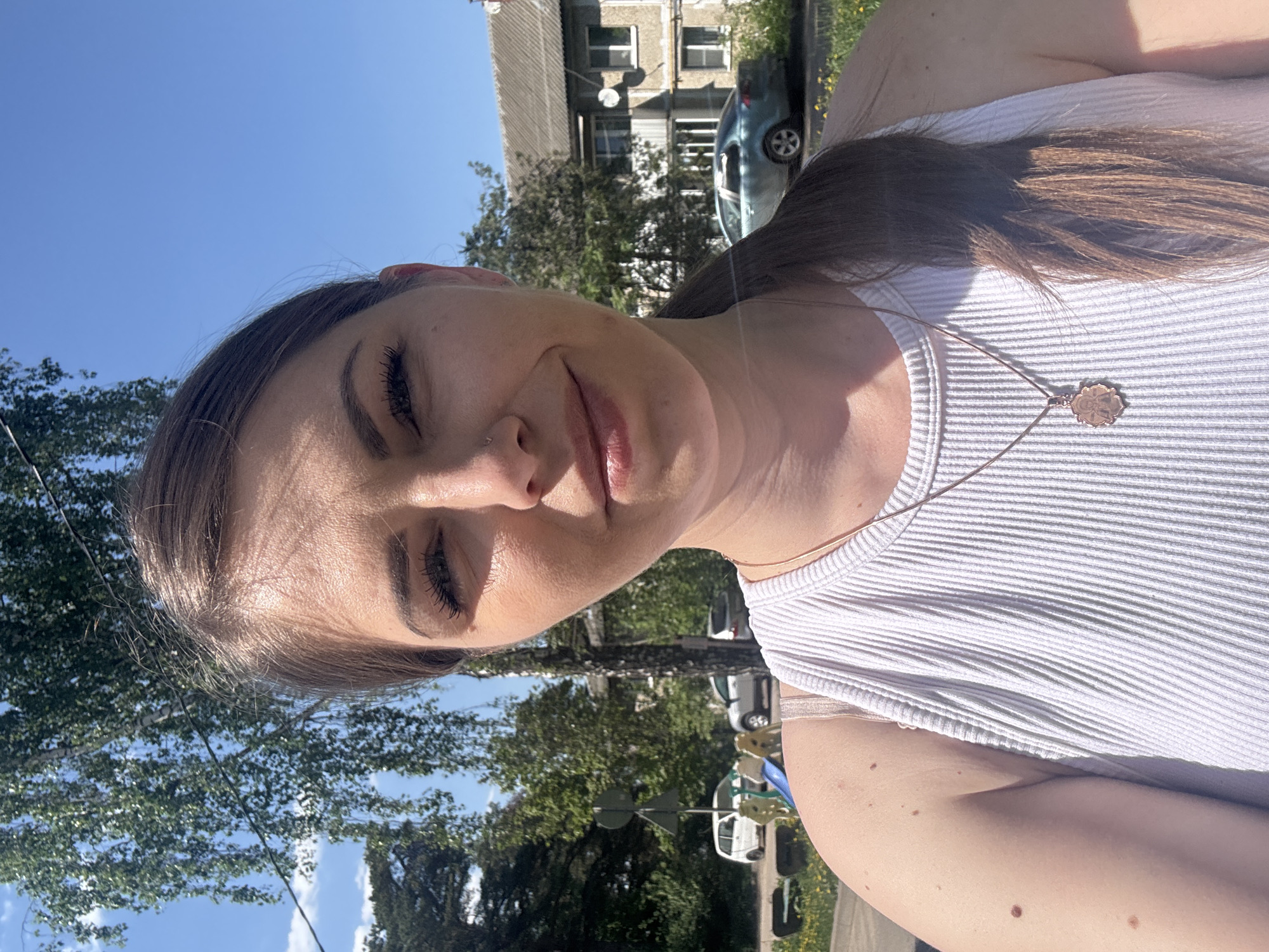 A young woman is captured in a selfie-style portrait, seemingly outdoors on a sunny day. She is wearing a white ribbed tank top and a delicate necklace with a round pendant. She has brown hair pulled back, fair skin, and is facing the camera with her eyes partially closed, perhaps due to the sunlight.
In the background, trees suggest an urban environment. Buildings are visible, including one that appears to be an older, Soviet-era apartment block with multiple windows and satellite dishes. Cars parked along the street further indicate a city location. Given the location, it is in the city of Razdolye, Russia.