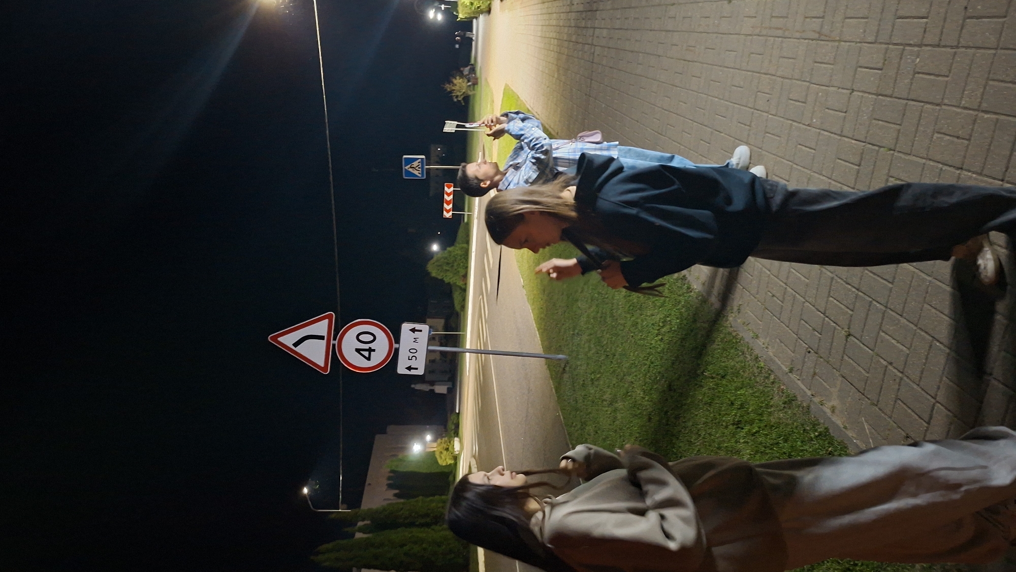 At night, three young people are standing on a sidewalk made of rectangular pavers, adjacent to a grassy area and a road. The person on the far left, a female, is facing left and appears to be wearing a light-colored hooded sweatshirt and pants. In the center, a female is facing right, gesturing with her hands and wearing a dark hooded sweatshirt and dark pants. The person on the right, appearing to be a male, is facing away from the camera, holding a smartphone to take a picture, and is wearing a light blue checkered shirt and jeans. A street light is visible in the background, illuminating the scene. Road signs are present, including a sign with a leftward curve arrow and a speed limit sign indicating 40, followed by a sign indicating "50 M" with upward arrows. Further down the sidewalk, a blue pedestrian crossing sign is visible. The environment suggests a street or road intersection within a town or city, possibly in Myadzyel, Belarus, given the presence of typical European road signage and pedestrian infrastructure.