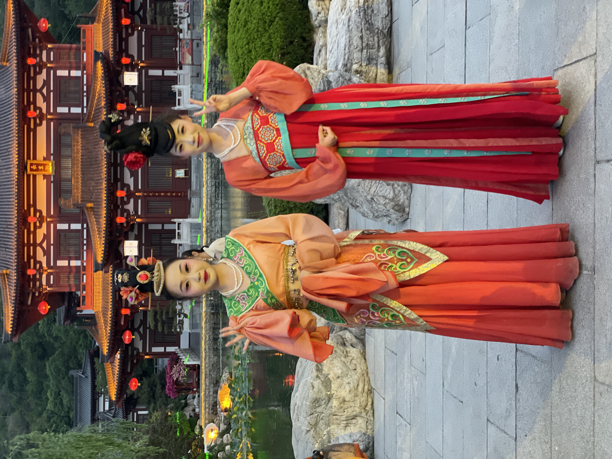 Two women in traditional Hanfu dresses pose for a photo in a park in Lintong District, China. The woman on the top wears a red Hanfu with green accents and a pearl necklace, while the woman on the bottom wears a peach-colored Hanfu with green accents and a pearl necklace. Both women are smiling and holding up peace signs. In the background, there is a traditional Chinese building with red lanterns, a pond with lily pads, and various plants and rocks. The ground is paved with gray bricks.