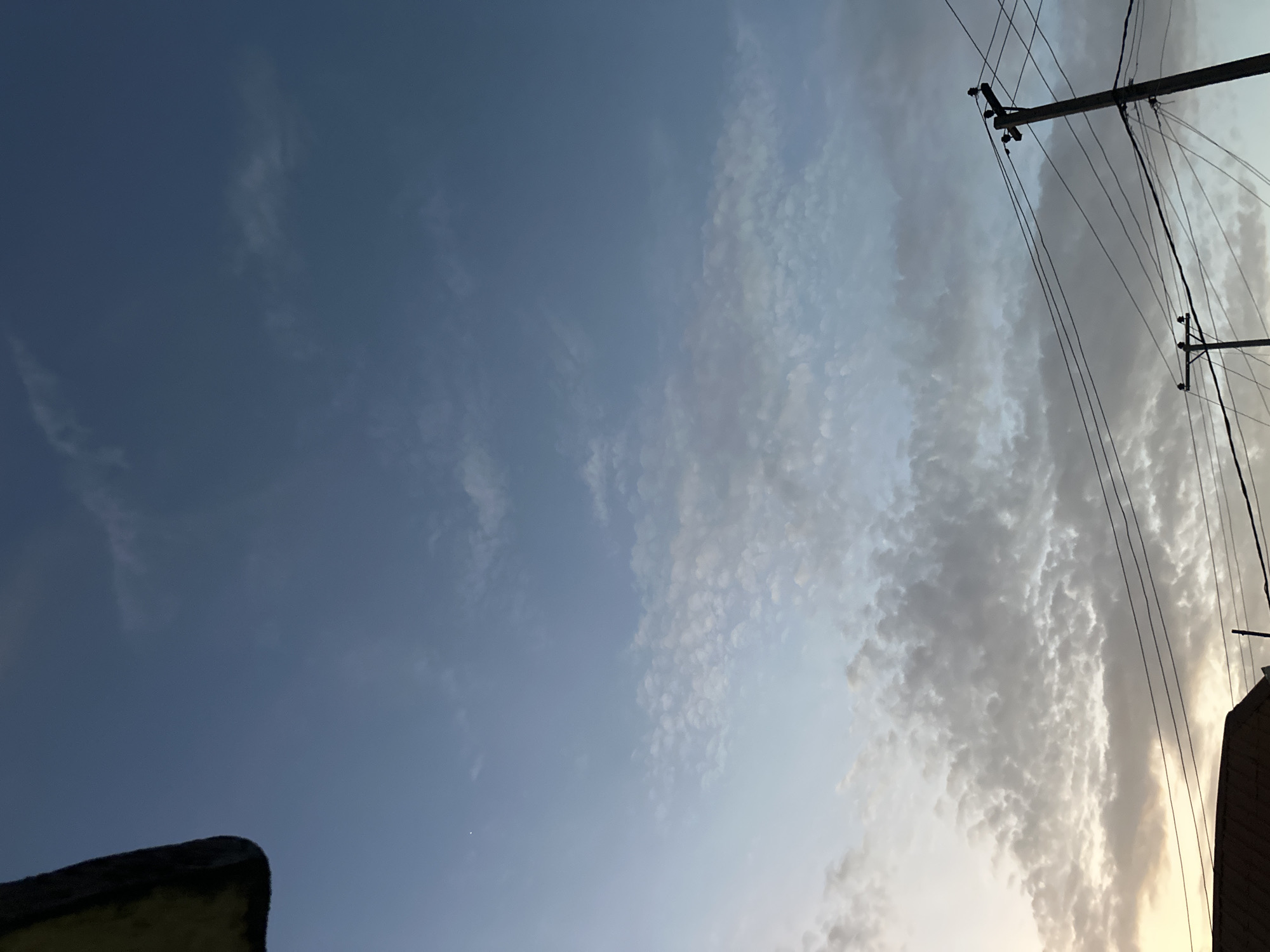 The image shows a sky with varying cloud cover. The left side of the image is mostly clear blue sky, while the right side is covered with textured, greyish-white clouds. Overhead power lines and utility poles are visible on the far right of the frame. In the bottom left corner, there is a dark, indistinct object that appears to be part of a building. In the bottom right, a building structure is seen.
The picture was taken in Jos, Nigeria. There is no specific evidence in the image that pinpoint the city of Jos, but it appears to be near sunset. It is a residential scene. The exact details of the location are not possible to determine. There are no people, subjects, actions, or interactions visible in the image.