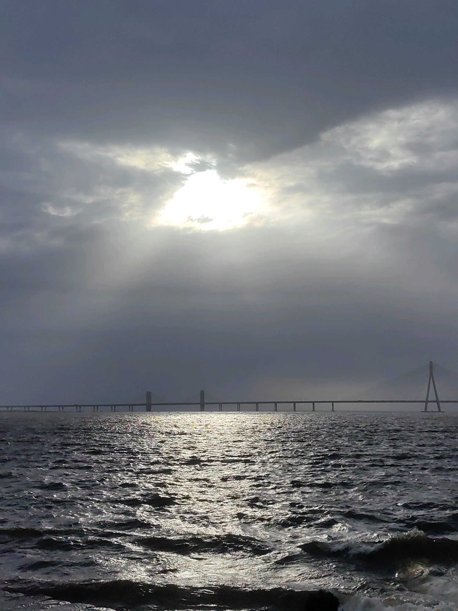 The image shows a wide view of the Bandra-Worli Sea Link bridge in Mumbai, India, during a cloudy day. The sun is partially obscured by clouds, creating a dramatic lighting effect on the water. The sea is choppy, with visible waves and reflections of the sun. The bridge, a long, slender structure with distinctive pylons, stretches across the frame, connecting two parts of the city over the Arabian Sea. The overall atmosphere is somber due to the overcast sky, but the sunlight breaking through adds a touch of hope. There are no people or vehicles visible on the bridge or in the water.