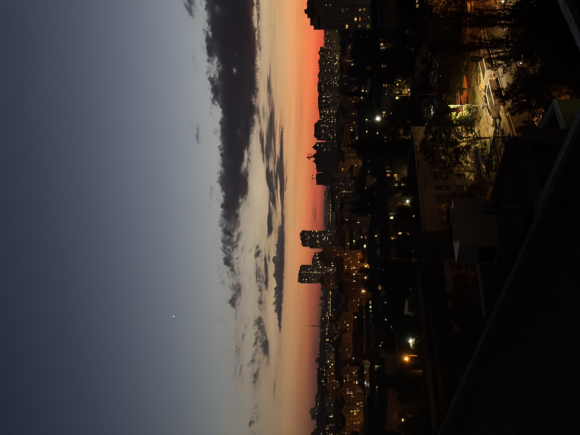 This is a high-angle, wide shot of a city skyline at dusk, likely in Kyiv, Ukraine. The sky transitions from a deep blue at the top to a vibrant orange and red near the horizon, indicating the sun has recently set. Several clouds are visible, some dark and textured, others lighter and wispy.
The city below is illuminated by numerous lights from apartment buildings and streetlights. Tall, modern buildings with many windows are prominent in the mid-ground, along with lower residential structures. Trees are scattered throughout the cityscape, some appearing to be in a park or courtyard area, their leaves silhouetted against the lights. In the foreground, the edge of a dark rooftop is visible, suggesting the vantage point is from a tall building. A single bright streetlight illuminates a small patch of ground in the lower right section of the image. No people are visible in the frame. The overall atmosphere is one of urban twilight.