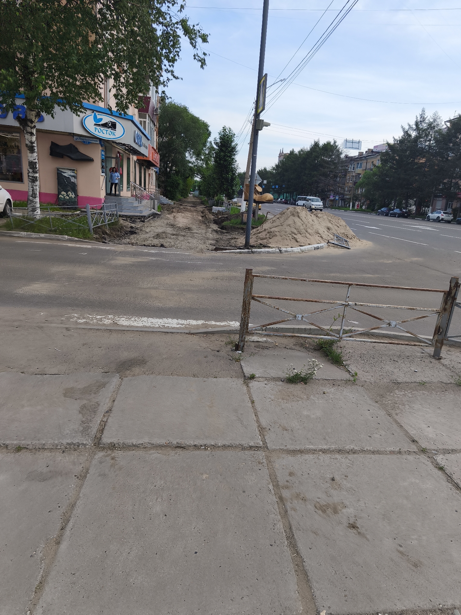 The image depicts a street scene in Komsomolsk-on-Amur, Russia, on a cloudy day. In the foreground, a concrete sidewalk is visible, with a rusted metal fence to the right. The sidewalk leads to a road where construction is underway. A large pile of sand is situated near the road, and excavation work is evident. A person in a blue shirt is standing near the entrance of a building on the left, which appears to be a commercial establishment with a sign that reads "ROSTOK". Further down the street, trees line the sidewalk, and several cars are parked along the road. The sky is overcast, and the overall impression is of an active construction zone within a city environment.