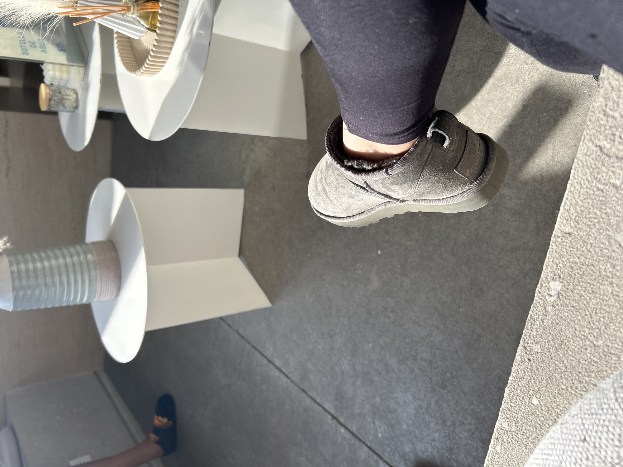 The image shows a first-person perspective, likely from an interior space with modern decor. A person is wearing dark leggings and a gray UGG slipper on their right foot. The flooring appears to be a combination of gray tiles and light-colored carpet or stone. In the periphery are elements of interior design, including white pedestal tables with cylindrical glass or plastic objects on top and other decorative items. The location seems to be a home interior with design-conscious elements.