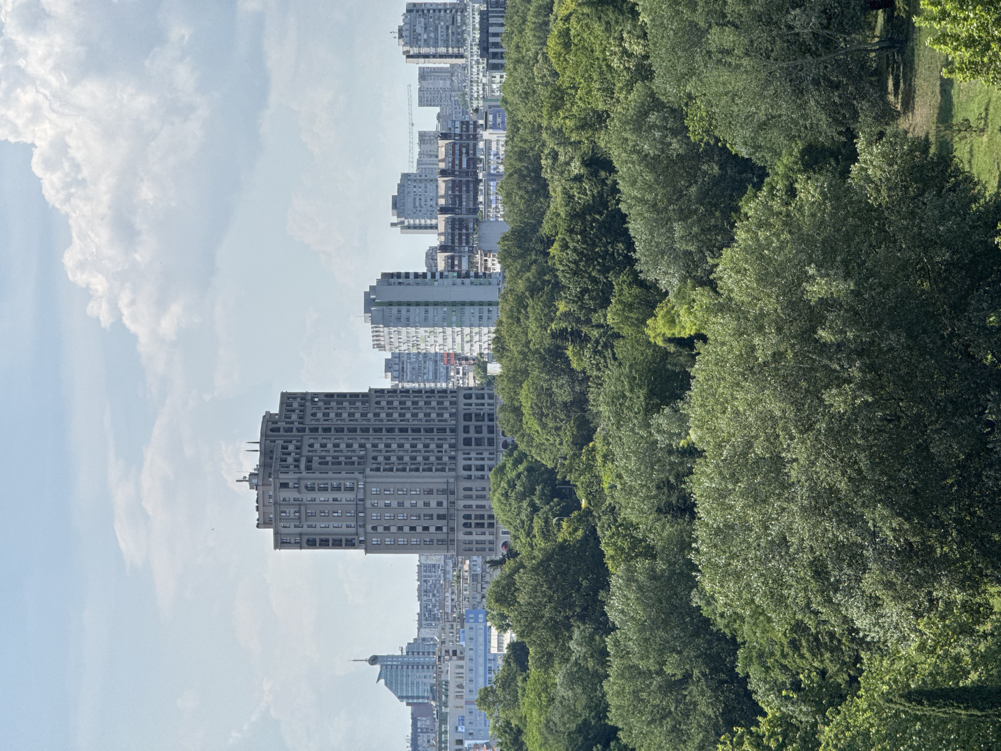 This image captures a panoramic view of the city of Burgas, Bulgaria, with a dense green forest occupying the foreground and mid-ground, and the city skyline in the background under a bright, partly cloudy sky.
The forest is characterized by a variety of deciduous trees, displaying lush green foliage. The canopy is thick, with varying shades of green suggesting different tree species and ages. Patches of sunlight filter through the leaves, creating a dappled effect on the ground below. In the far right mid-ground, a small grassy clearing is visible amidst the trees.
The city skyline features a mix of architectural styles, dominated by apartment buildings and high-rise structures. Prominently, a tall, ornate building with a patterned facade and arched windows stands out. To its left, a modern skyscraper with a distinctive curved top is visible. Further to the right, a cluster of residential blocks, some with colored balconies, are interspersed with other office buildings and structures. A construction crane is visible in the far distance, suggesting ongoing development.
The sky is a light blue, with scattered white cumulus clouds, indicating fair weather conditions. The overall scene presents a harmonious blend of urban development and natural landscape, likely depicting the edge of Burgas where residential areas meet a significant green space.