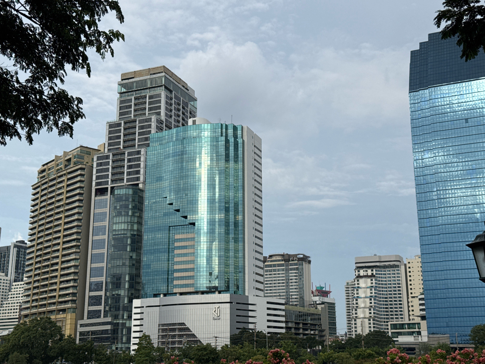 This is a photograph of a cityscape in Bangkok, Thailand, likely taken from a lower elevation, possibly a park or garden area. In the foreground, a dark leafy tree branch partially obscures the left side of the frame. The dominant features of the image are several modern high-rise buildings.
To the left, a tall, light-colored building with numerous horizontal window sections is visible. Adjacent to it, and slightly behind, is another tall building with a predominantly glass facade, reflecting the sky. In the center of the image, a striking building with a turquoise-green glass exterior stands prominently. This building has a unique architectural design with angled sections and setbacks. It appears to be a commercial building or a hotel. Below this glass tower, a lighter-colored, multi-story structure with horizontal louvers or window accents is visible.
Further to the right, another very tall building with a blue glass facade dominates the right side of the image. This building has a segmented top. Behind the central glass tower, other buildings of varying heights and architectural styles can be seen, including a white building with a grid-like facade and a darker, more industrial-looking structure in the distance. The sky is overcast with scattered clouds. The lower parts of the image show lush green trees and some pink flowering bushes, suggesting a green space within the urban environment. The overall impression is one of a modern, bustling city with contemporary architecture.
