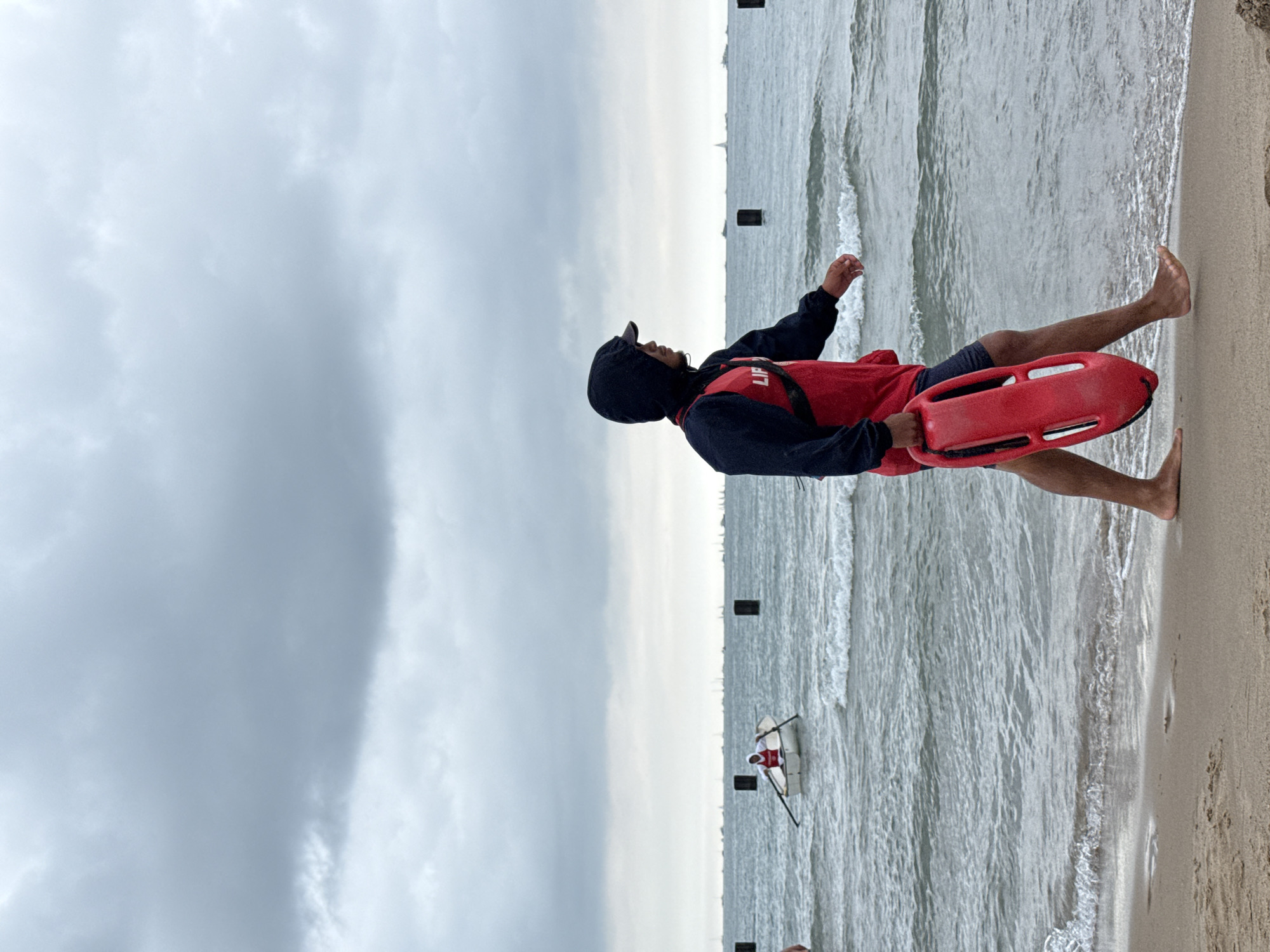 A lifeguard wearing a red vest over a dark hoodie and shorts walks along the sandy shore of Lake Michigan. The lifeguard carries a red rescue buoy in their right hand. In the background, several sailboats are visible on the choppy, gray water under an overcast sky. A small white boat with a person rowing is also in the water, further from the shore. Dark posts are visible in the water at intervals. The scene suggests a cloudy day at a Chicago beach.