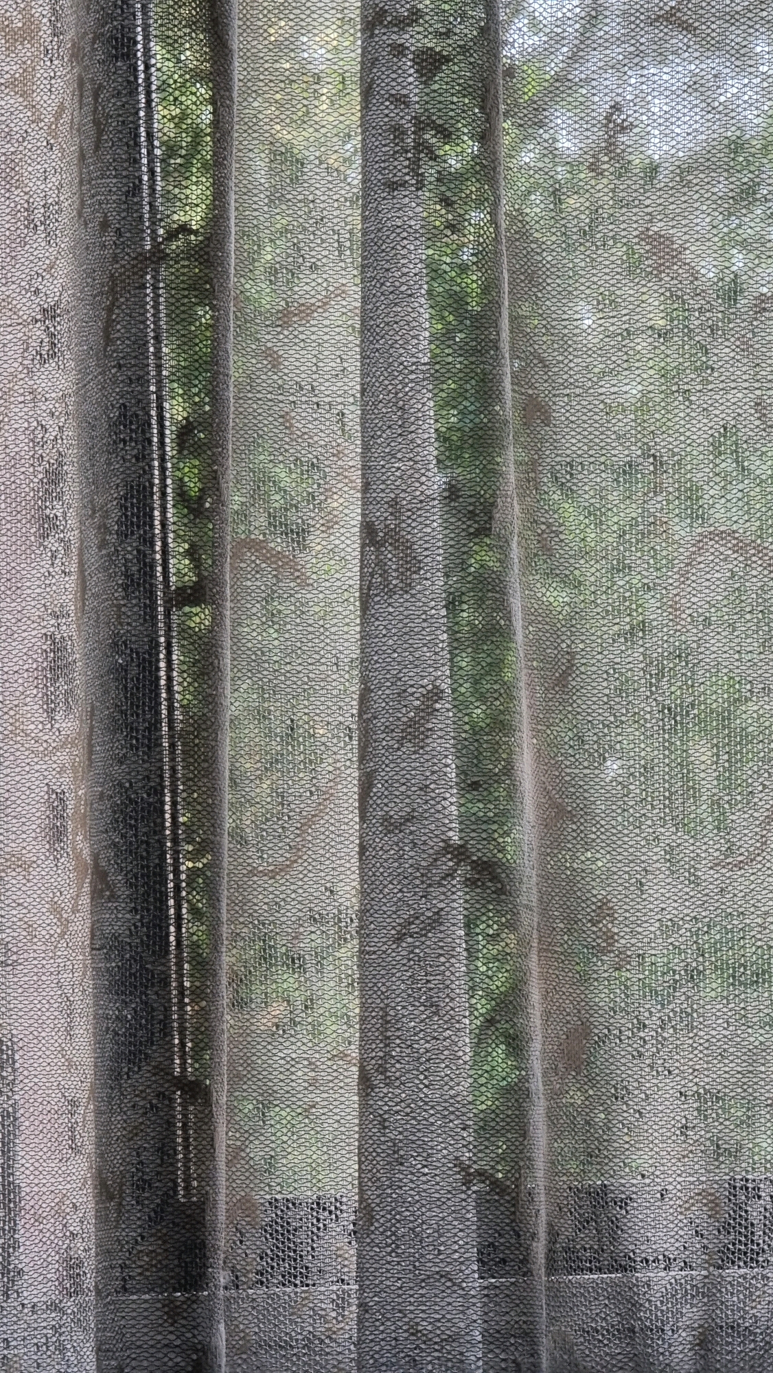 The image shows a close-up view of sheer curtains hanging in front of a window. The curtains have a textured, patterned weave, with vertical folds creating shadows and depth. Through the semi-transparent fabric, glimpses of green foliage are visible, suggesting an outdoor scene with trees or plants. The lighting is diffused, creating a soft, somewhat muted atmosphere. The scene appears to be an interior shot looking outwards, with the curtains acting as a visual filter. No people, specific objects, or actions are discernible within the frame. The location in Perondi, Albania is not visually represented in the image itself.