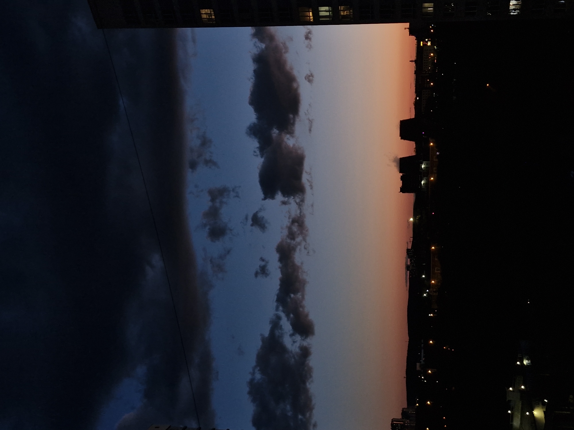 This vertically oriented image captures a dramatic twilight or dawn scene over the city of Yekaterinburg, Russia. The predominant subject is the sky, which exhibits a striking gradient of colors and cloud formations.
The left side of the frame is dominated by a very dark, deep blue-grey sky with heavy, obscure clouds, suggesting an area already well into darkness or intense weather. Moving towards the right across the frame, the sky transitions to a lighter, clearer blue, revealing more distinct dark grey cumulus clouds of varying shapes and sizes. Further to the right, the sky brightens considerably, displaying soft orange and pink hues, indicative of the lingering light from a setting or rising sun. A thin, dark wire or cable stretches horizontally across the sky, bisecting the cloudscape.
At the very top edge of the image, a dark building silhouette with a few illuminated windows is visible, suggesting the perspective is from a high vantage point within an urban structure. Along the right side, forming what would be the horizon if the image were rotated, is the dark silhouette of a cityscape. Numerous small, bright lights, likely from streetlights and buildings, punctuate the dark urban sprawl, beginning to illuminate the evening or morning. The bottom right corner of the image is completely dark.
The time of day is clearly twilight, characterized by the dynamic interplay of dark clouds, transitioning sky colors, and the onset of artificial city lights. No people or visible text are discernible in the image.