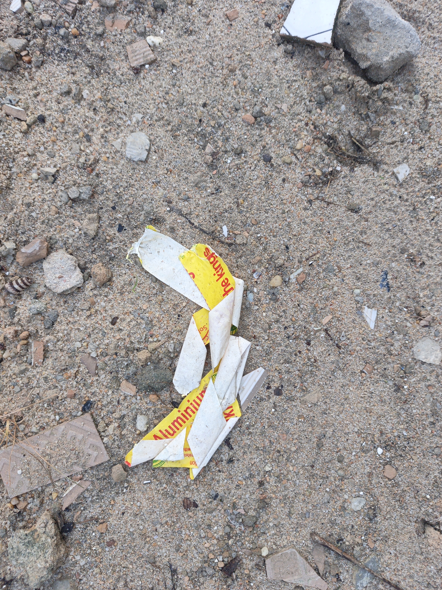 The scene is a close-up, ground-level view of sandy, pebbled terrain littered with debris. Visible detritus includes broken tile fragments, pieces of brick or concrete, and torn pieces of packaging with yellow and white coloration. The packaging appears to be partially legible, with the words "he Kings" and "Aluminium" visible. The location appears to be urban, with the presence of construction debris and packaging indicative of human activity. The specific city and other contextual details are unidentifiable from the image.