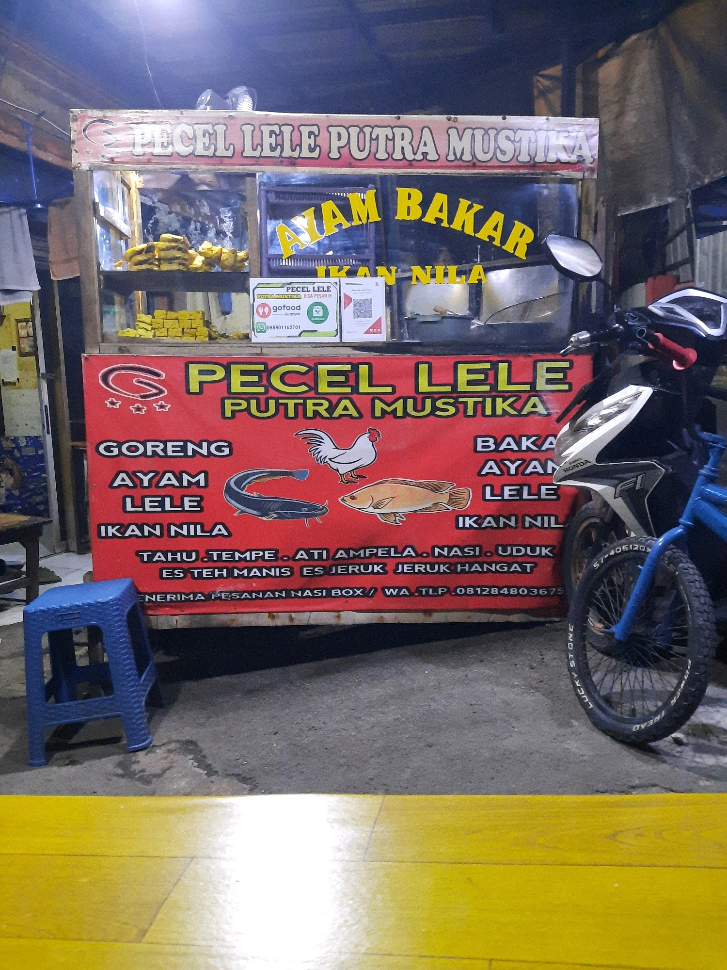 The image depicts a food stall at night in East Jakarta, Indonesia, specifically at the "PECEL LELE PUTRA MUSTIKA" stall. The stall is a mobile cart with a red banner displaying the menu items and the business name. Visible menu items include "AYAM BAKAR" (grilled chicken), "LELE GORENG" (fried catfish), "AYAM GORENG" (fried chicken), "IKAN NILA" (tilapia fish), "TAHU TEMPE" (tofu and tempeh), "ATI AMPELA" (chicken gizzards and proventrails), "NASI UDUK" (coconut rice), "ES TEH MANIS" (sweet iced tea), and "ES JERUK HANGAT" (warm orange juice). The stall also advertises accepting orders via GoFood and provides a phone number for inquiries and orders: 081284803675. A blue plastic stool is placed to the left of the stall, and a motorcycle is parked to the right, partially obscuring the stall's display. A blue bicycle is also visible on the right side of the image. The background is dark, suggesting it is nighttime, and shows some structural elements of a market or street vendor area. The ground is a combination of asphalt and a yellow painted surface.