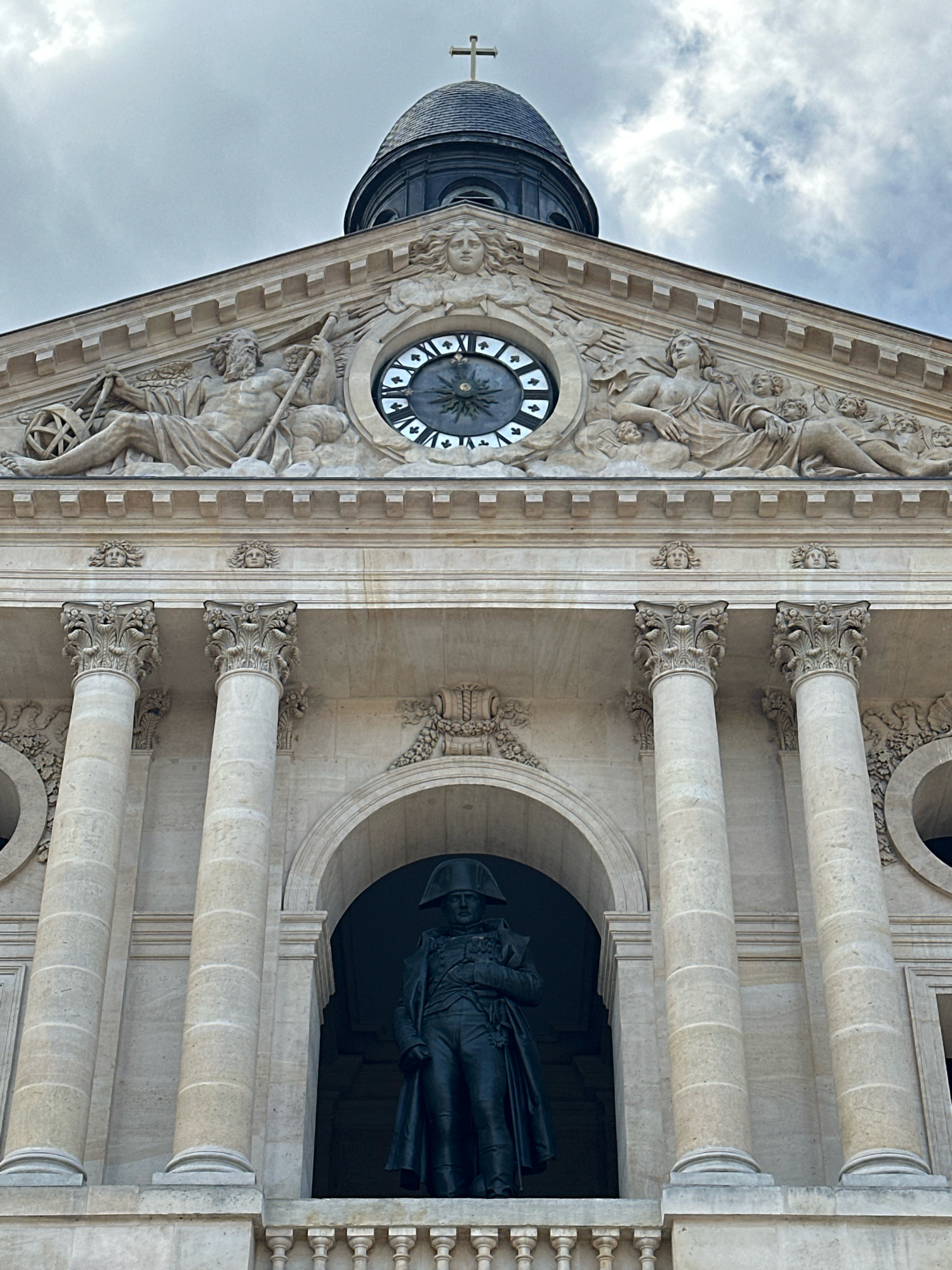 The image depicts the facade of the Hôtel des Invalides in Paris, France, under a cloudy sky. At the apex of the building's pediment, a cross tops a dark, domed cupola. The pediment is adorned with intricate bas-relief sculptures. A large, classical clock face with Roman numerals is centrally placed within the pediment. Flanking the clock are sculpted figures: on the left, a bearded male figure reclines with a globe-like object and a staff; on the right, a female figure reclines with several cherubic figures. Below the pediment, a row of Corinthian columns supports a heavy entablature. Between two of these columns, within a recessed arched niche, stands a dark bronze statue of Napoleon Bonaparte. The statue portrays Napoleon in military attire, wearing a bicorne hat and a long coat, with his hands clasped in front of him. The architectural style is classical, with elaborate detailing on the columns and facade.