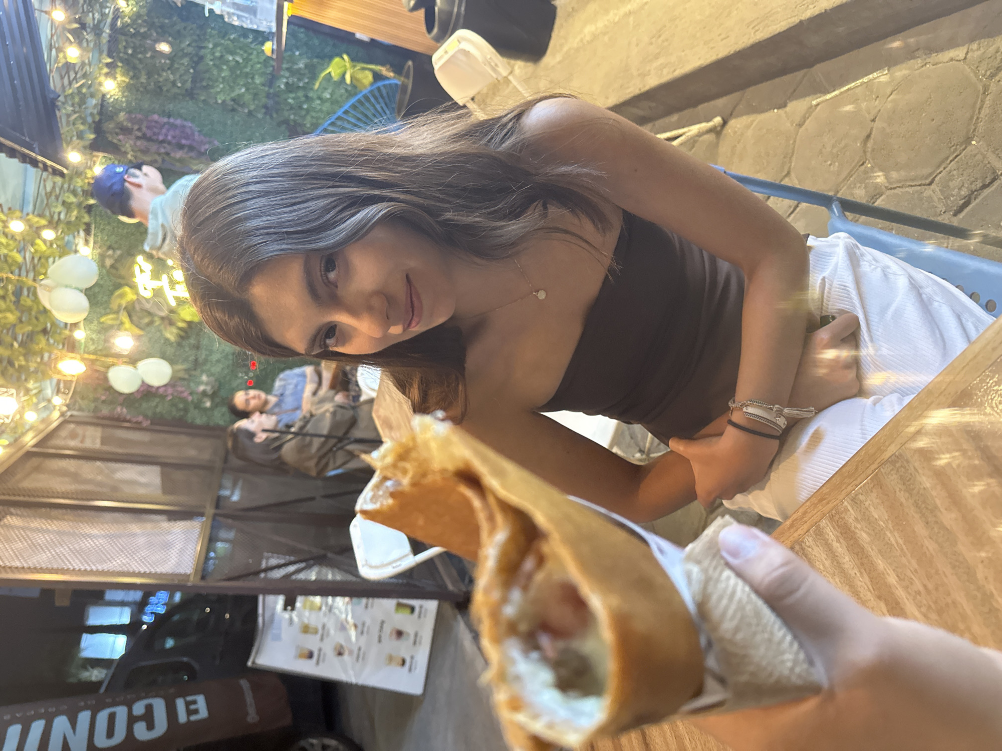 The image captures a woman seated outdoors, holding a crepe in her hand, likely ready to eat it. She has long brown hair and is wearing a brown tube top. In the background, other people are seated, and fairy lights adorn the surrounding structures. A menu board can be seen next to a food stall or truck. The presence of a restaurant menu and outdoor seating suggests this is a public space in Puebla, Mexico. The woman is looking at the camera and smiling.