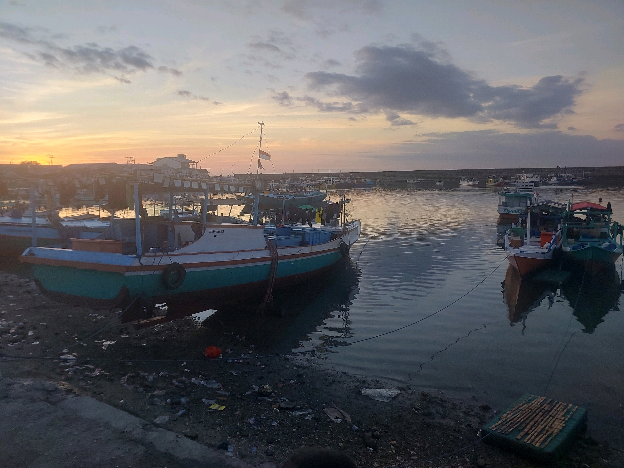 This image depicts a harbor scene at dusk in the city of Kupang, Indonesia. Several fishing boats are moored near a shoreline that is littered with trash. In the background, buildings and structures line the coast, and the sky is illuminated by the warm glow of the setting sun, with scattered clouds adding texture to the horizon. The water is calm, reflecting the sky and the boats. The overall impression is one of a working harbor at the end of the day.